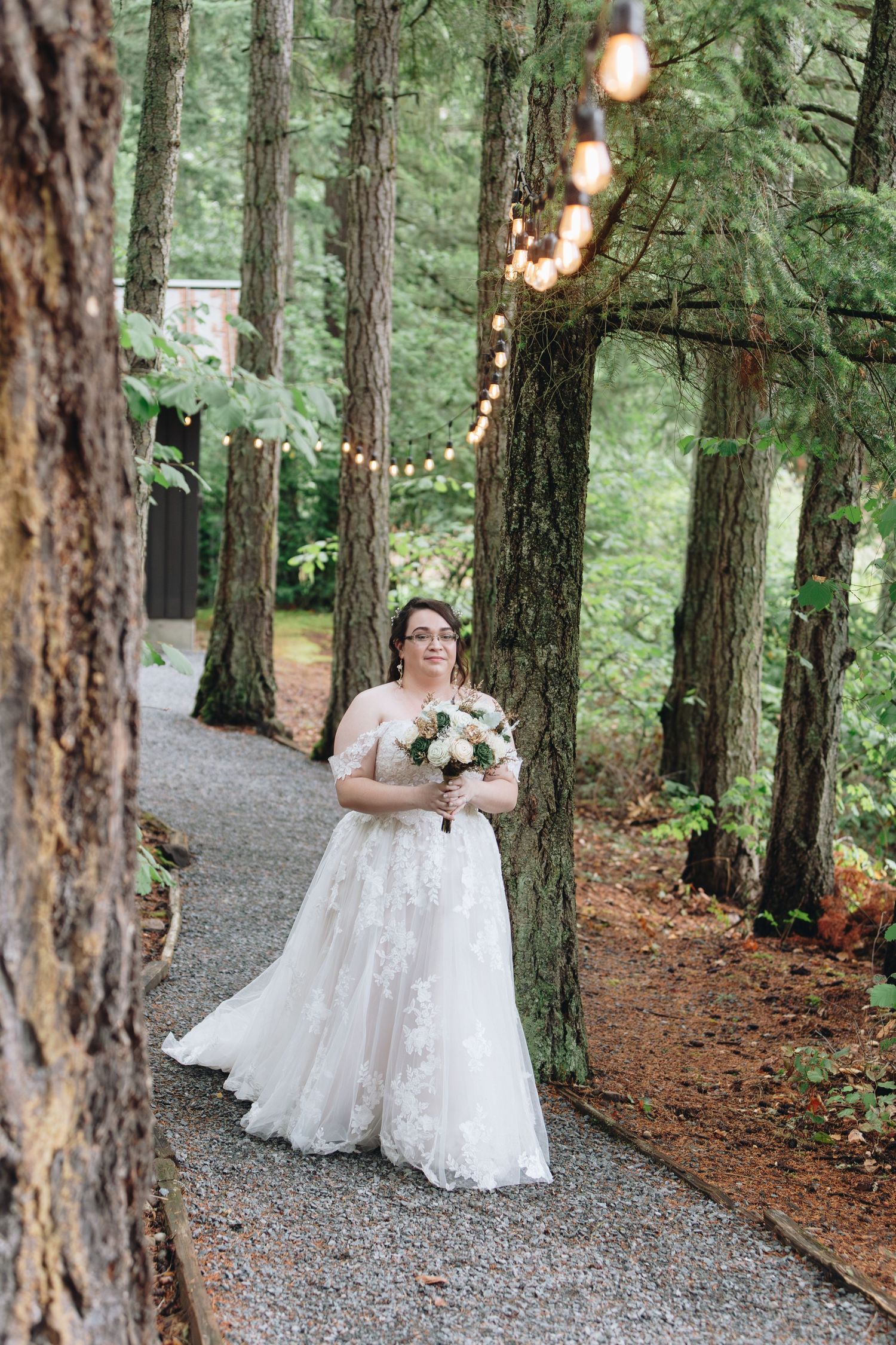Bride in lace wedding gown holding bouquet on forest path with string lights among tall trees.