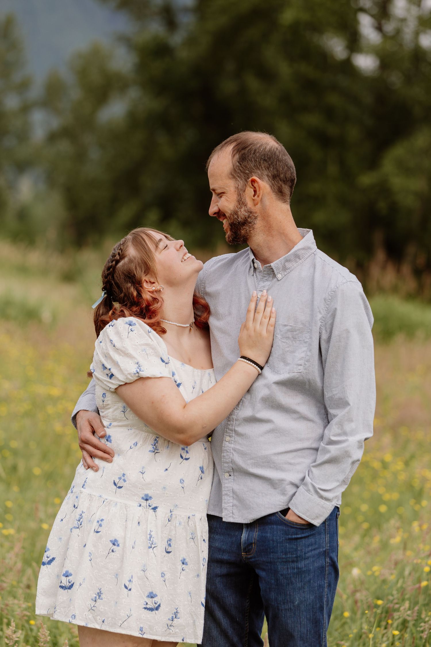 Couple embracing and smiling at each other in a sunny wildflower meadow, woman in floral dress, man in gray shirt.