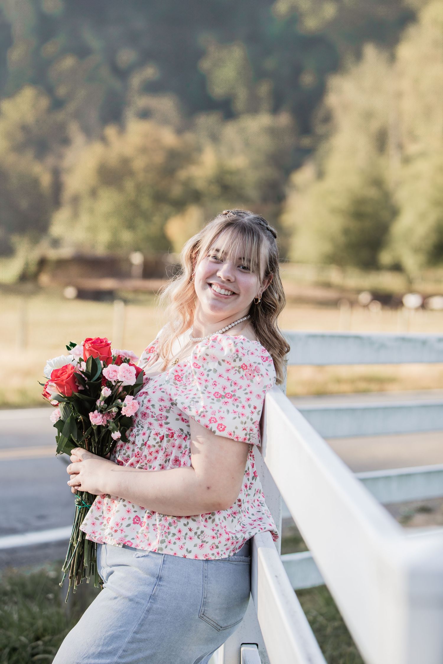 Smiling young woman holding pink roses in a floral top leaning on a white fence.