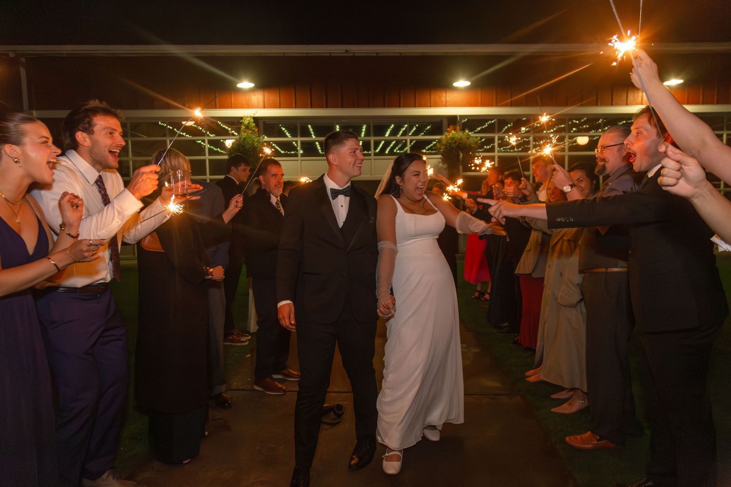 Bride and groom walk through a sparkler tunnel surrounded by cheering wedding guests at night.