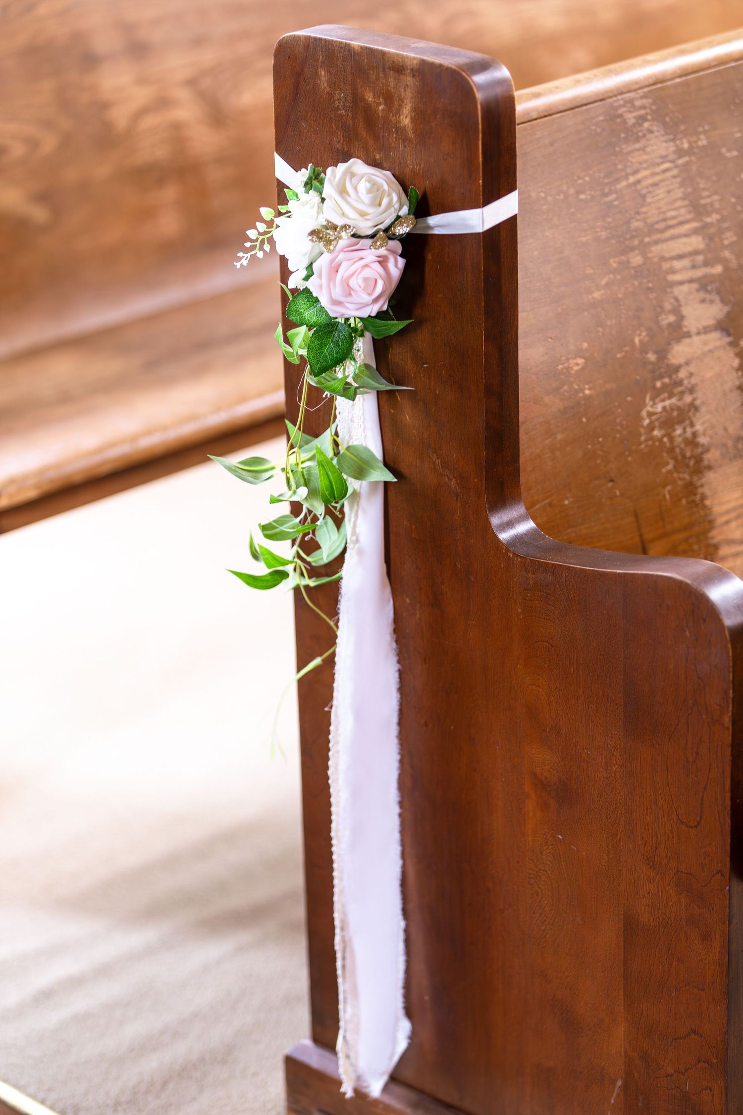 Elegant wedding pew decorated with pink and white roses, greenery, and white ribbon in a church.