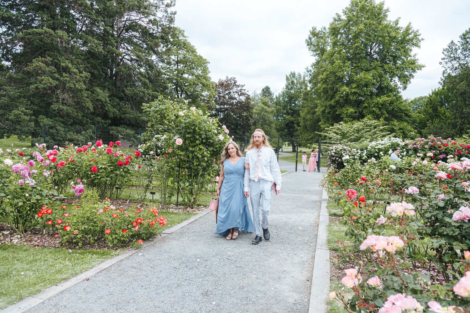 Two women walking along a flower-lined garden path surrounded by colorful roses.