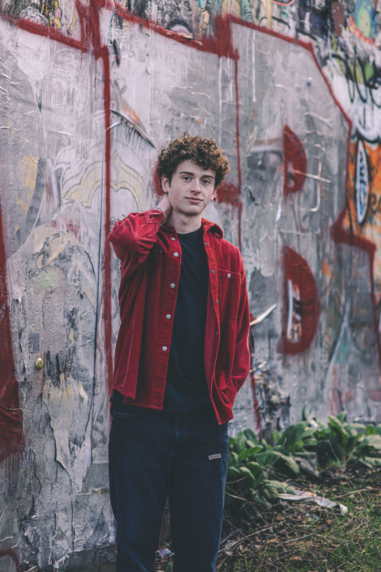 Teen boy in red jacket posing confidently against urban graffiti-covered wall outdoors.