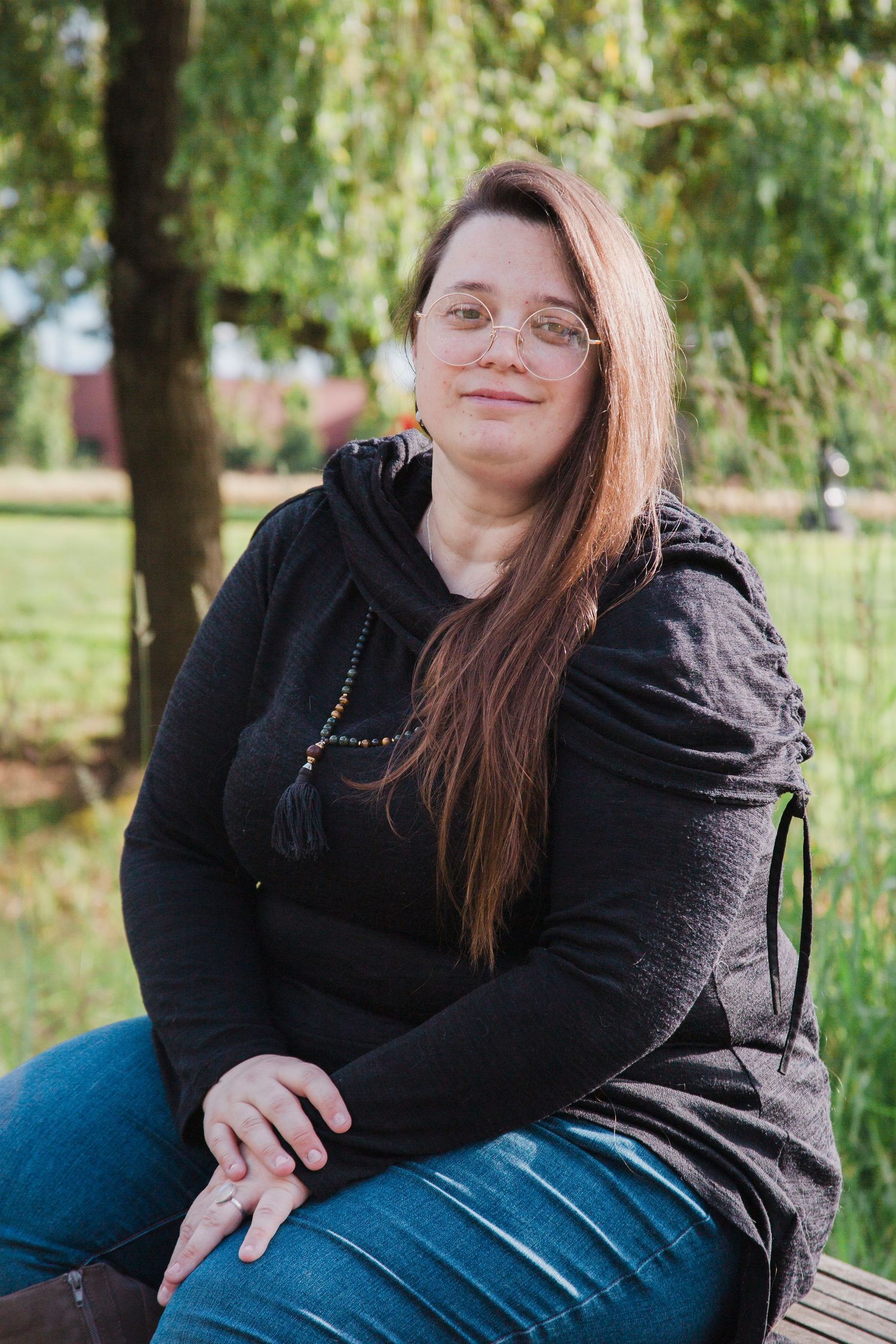 Smiling woman with long red hair and glasses sitting on bench in sunny green park.