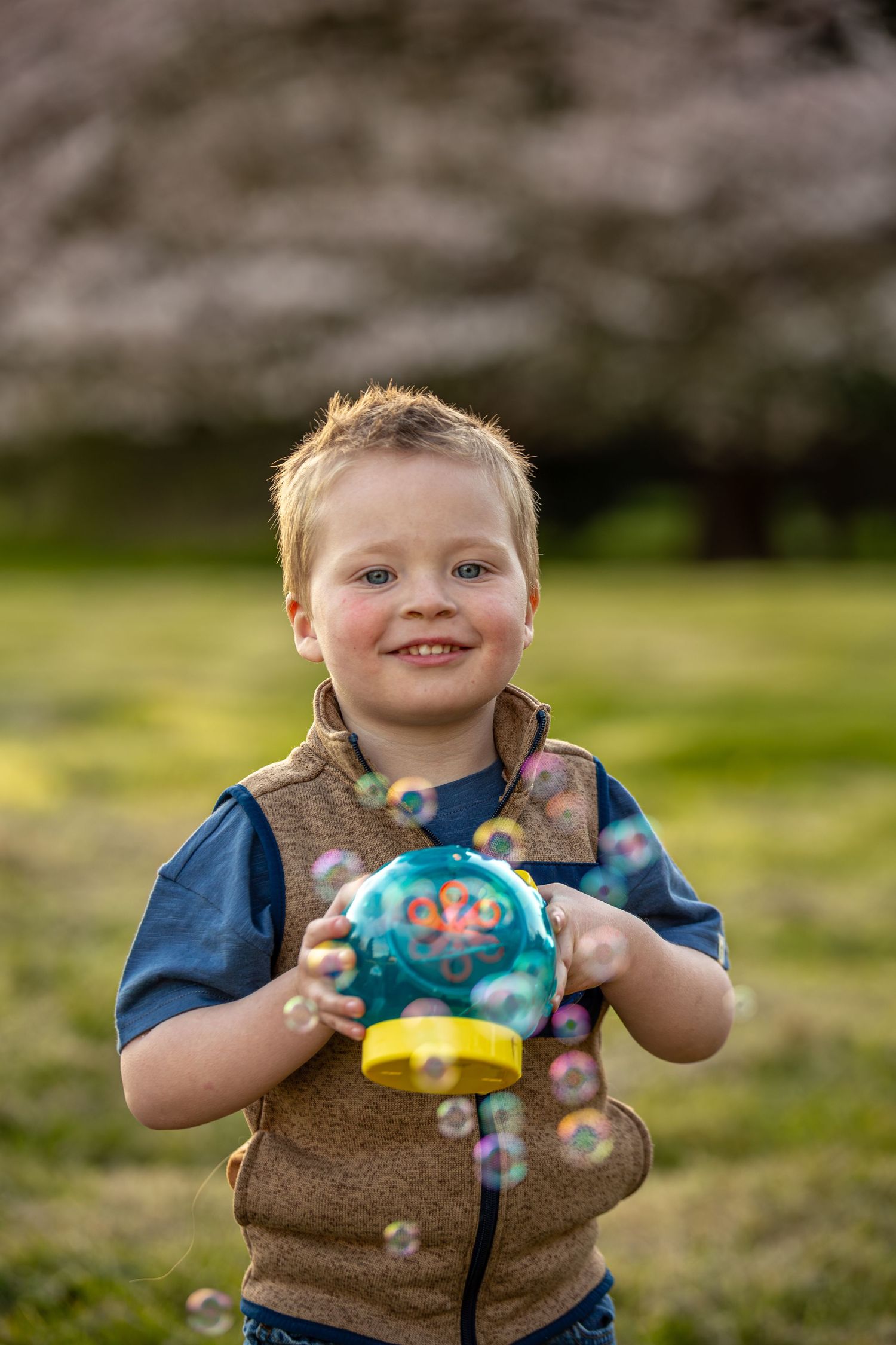 Happy young boy holding a colorful bubble machine surrounded by bubbles with cherry blossoms behind him.