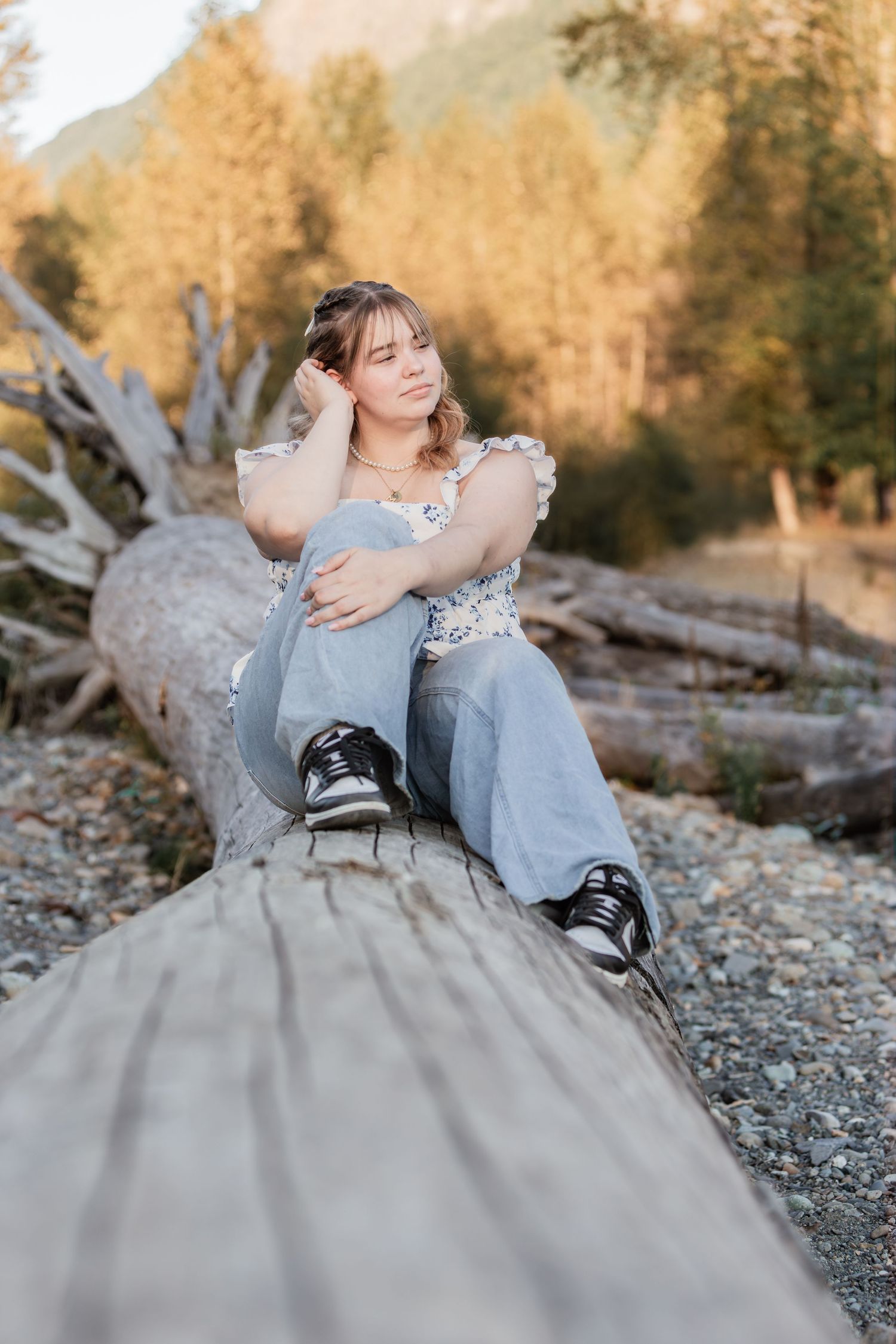 Young woman in floral top and jeans sitting on a fallen log in a scenic outdoor setting with trees and mountains.