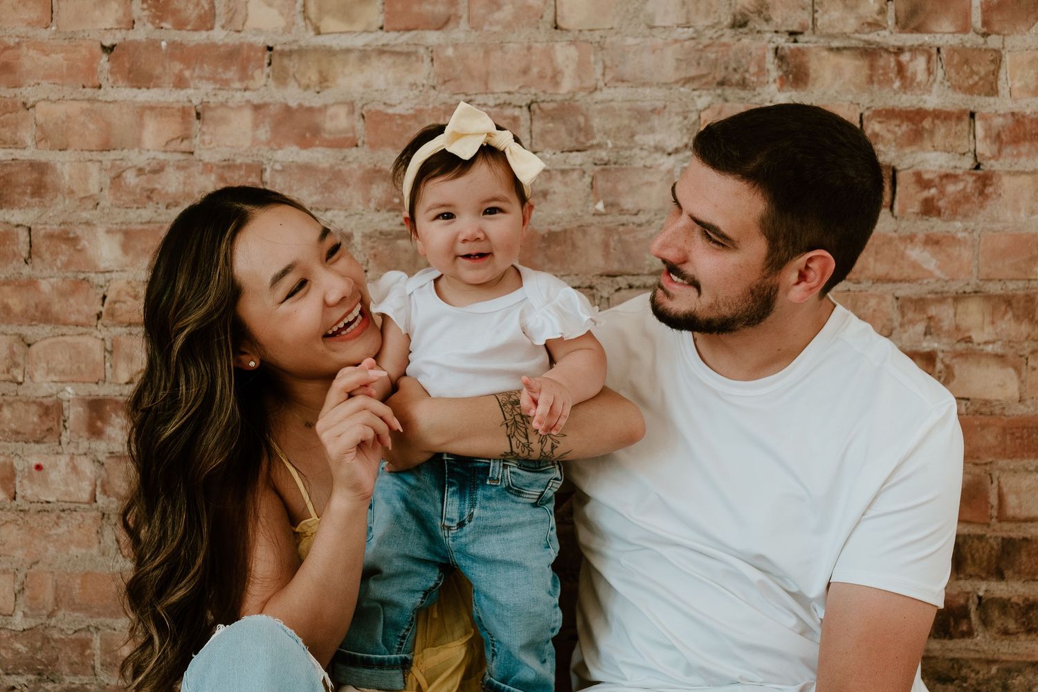 Young family with baby smiling together in front of rustic brick wall