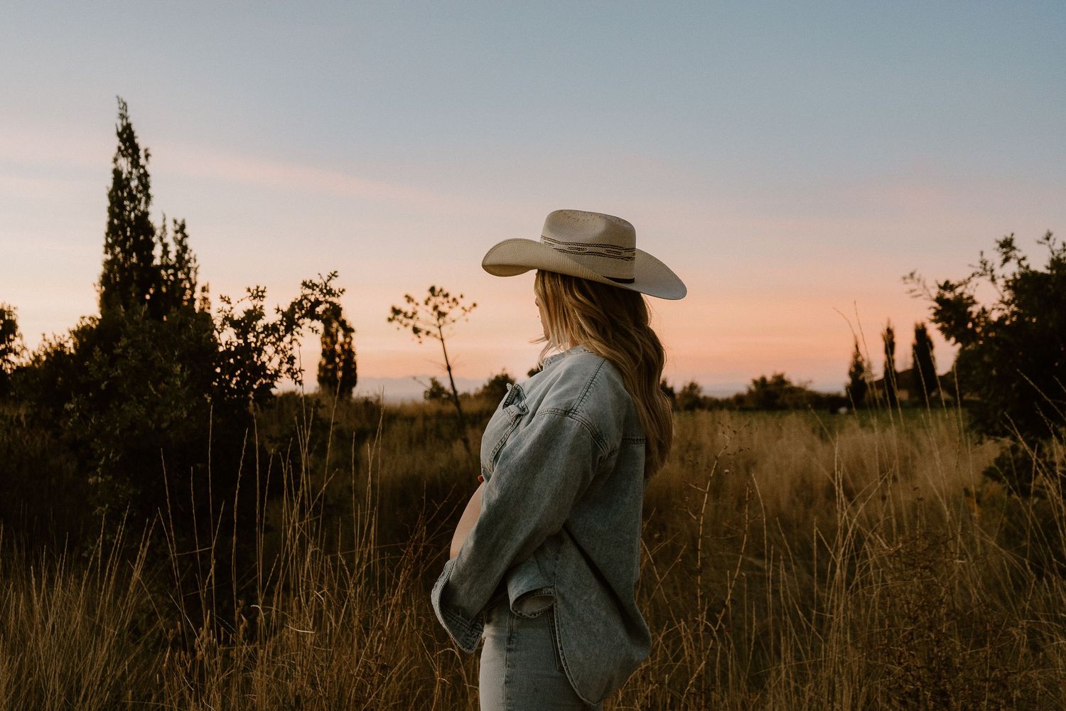 Woman in cowboy hat standing in field at sunset during Utah maternity portrait session