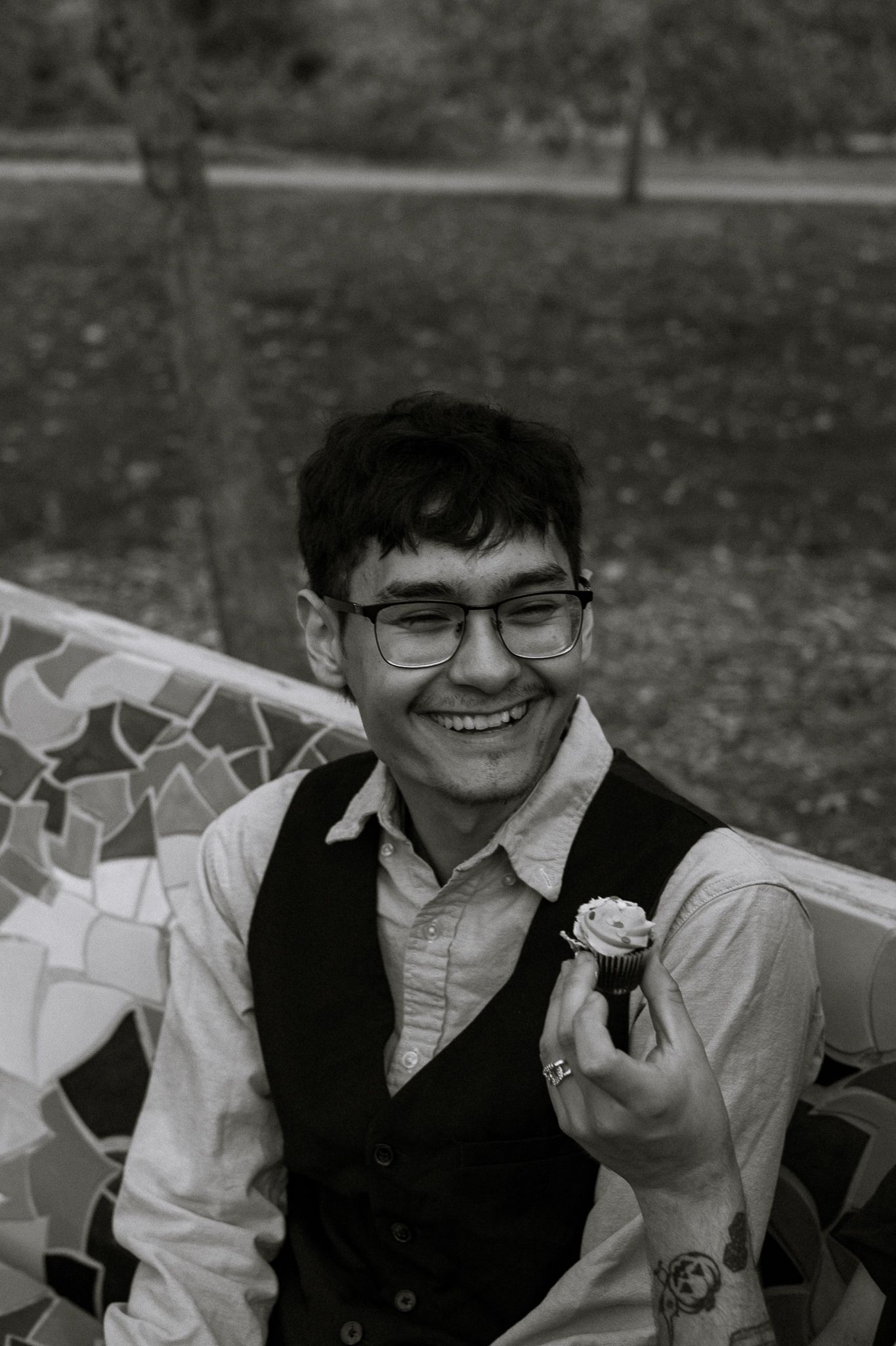 Black and white portrait of a smiling young man in a vest holding a boutonniere outdoors.