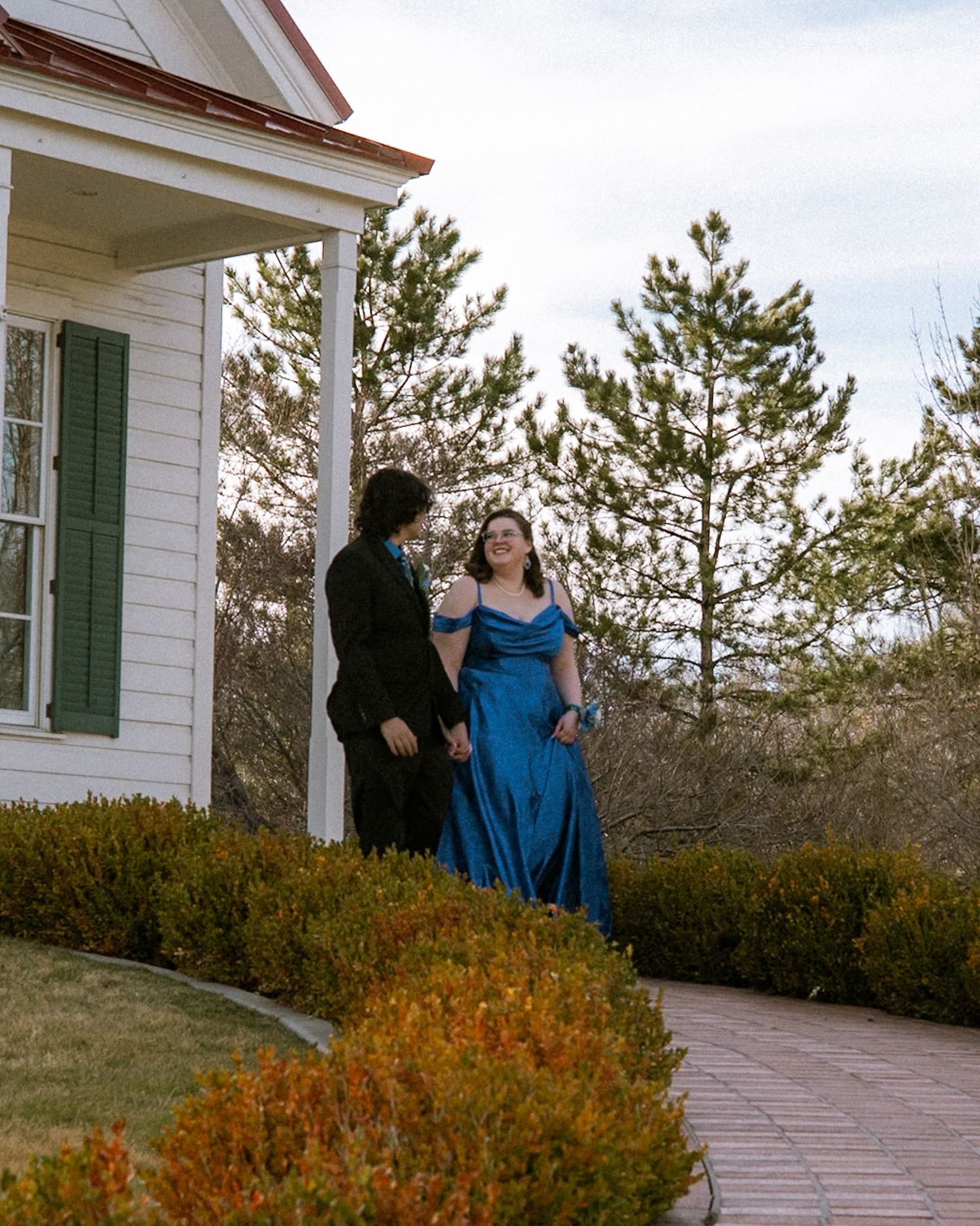 A couple in formal wear walks along a garden path beside a white building with pine trees.