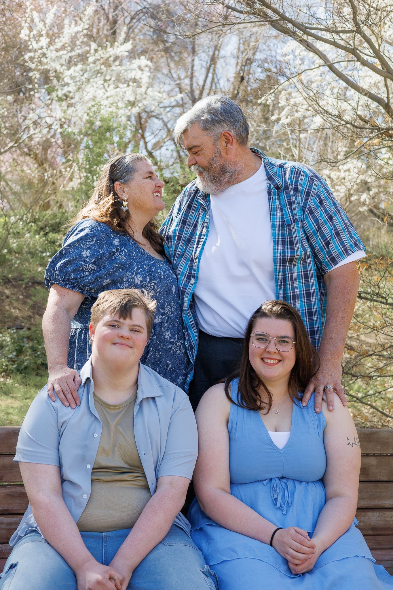 Happy family of four posing outdoors in spring, parents standing behind two adult children seated on a bench.