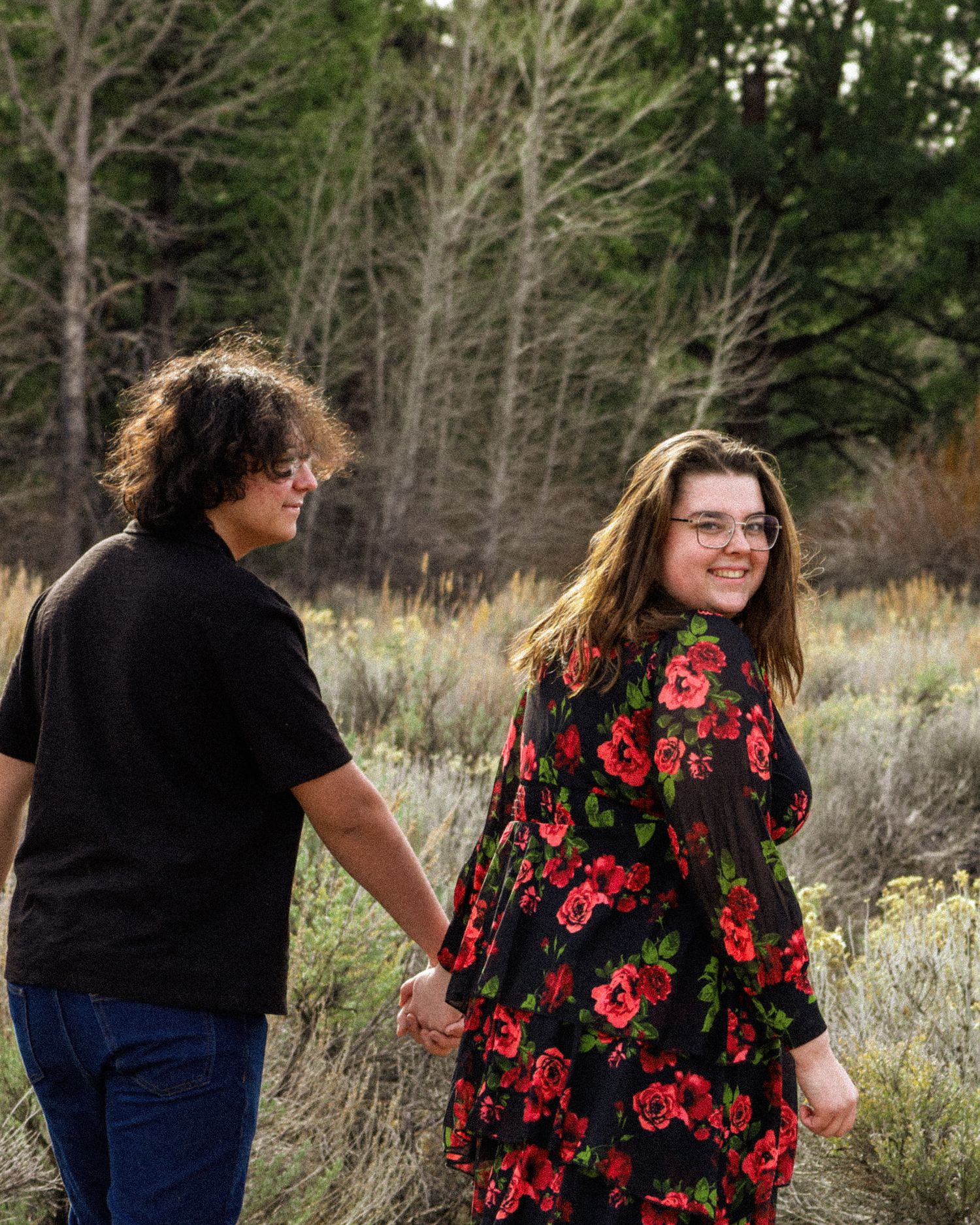 Young couple holding hands walking through a natural meadow, woman in floral dress looking back smiling.