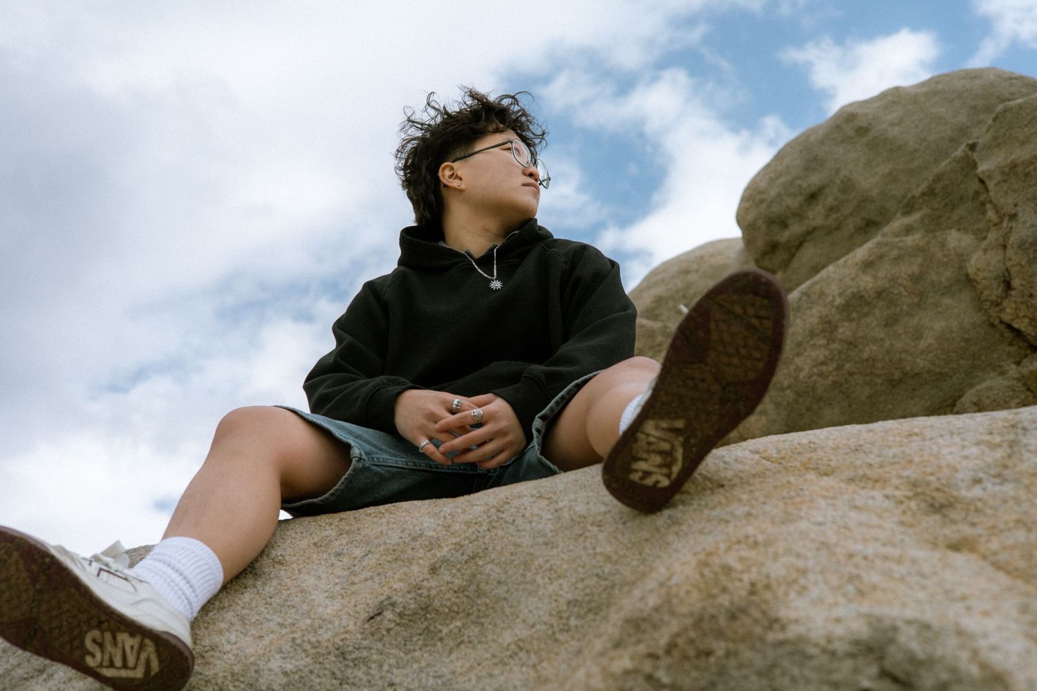 Young person in black hoodie sitting on boulder, looking up at cloudy sky from low angle.