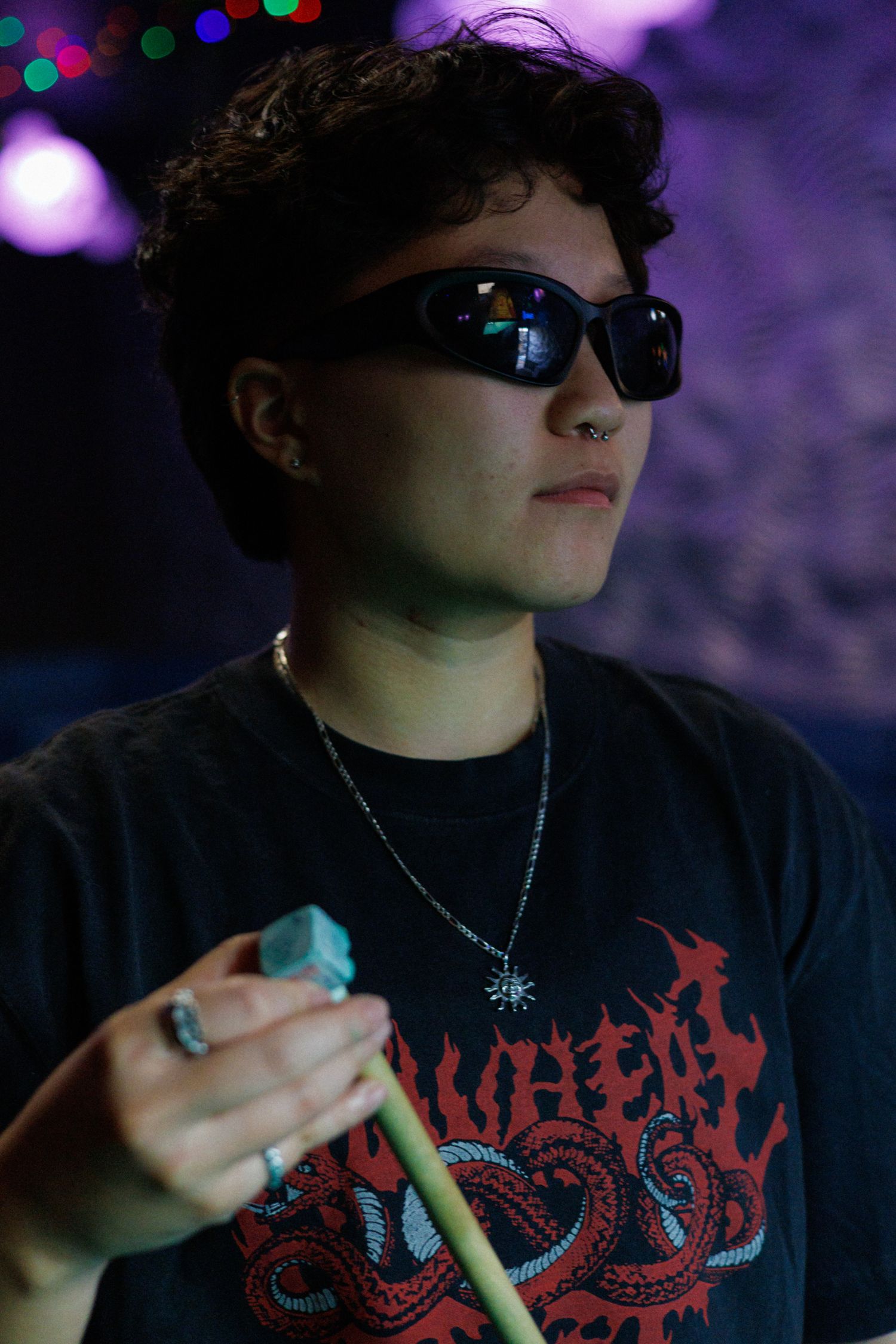 Young person wearing sunglasses and a black metal band tee holds a pool cue at a dimly lit venue.