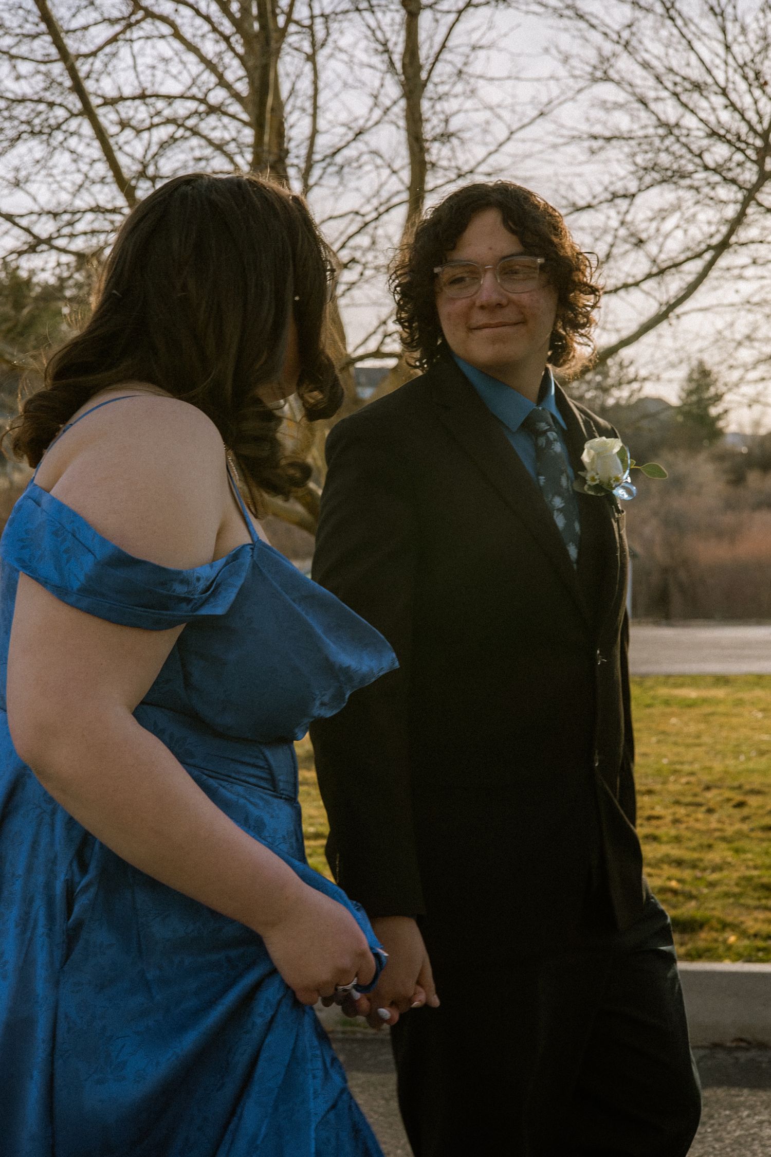 Young man in black suit smiles back at date in blue satin prom dress while holding hands outdoors.