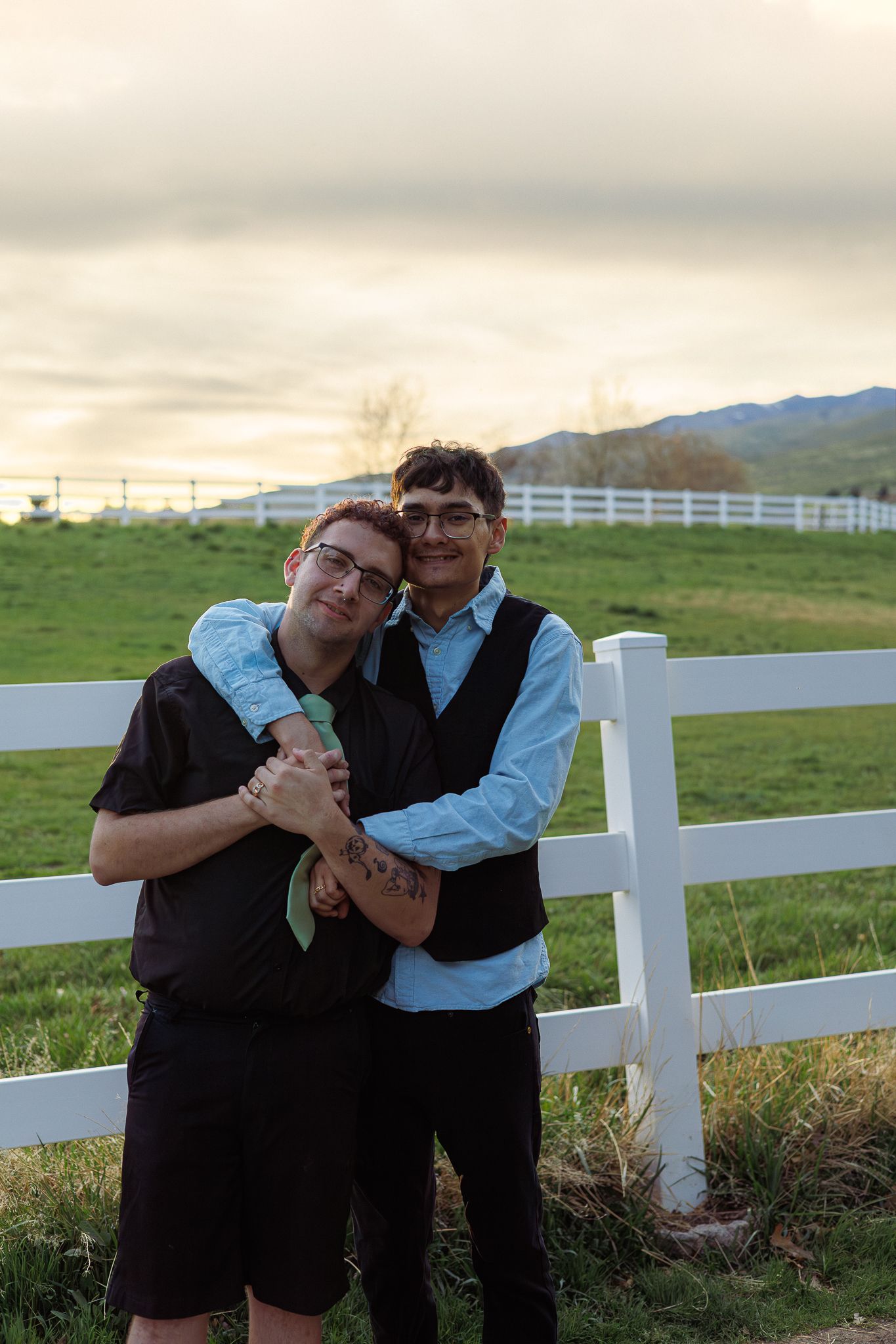 Two men embrace warmly by a white fence at sunset with green fields and mountains behind.