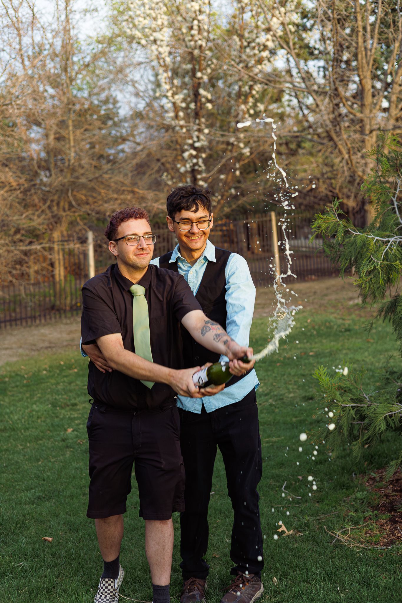 Two men in formal attire celebrate outdoors by popping a champagne bottle in a garden.