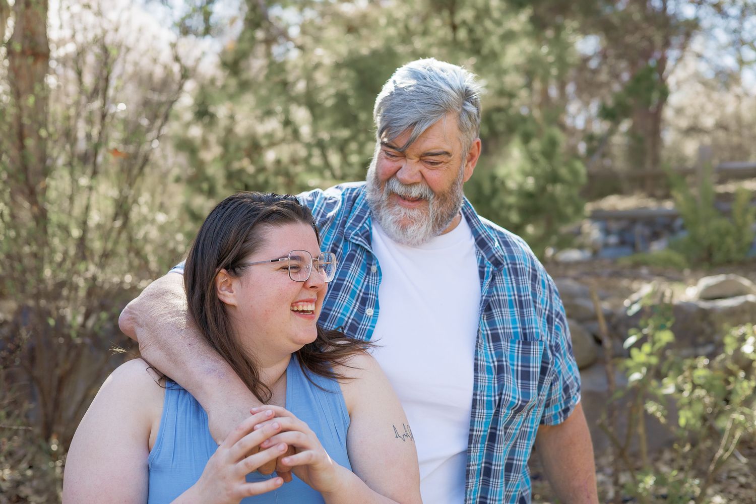 Older man with gray beard and young woman with glasses smiling together in a sunny outdoor setting.