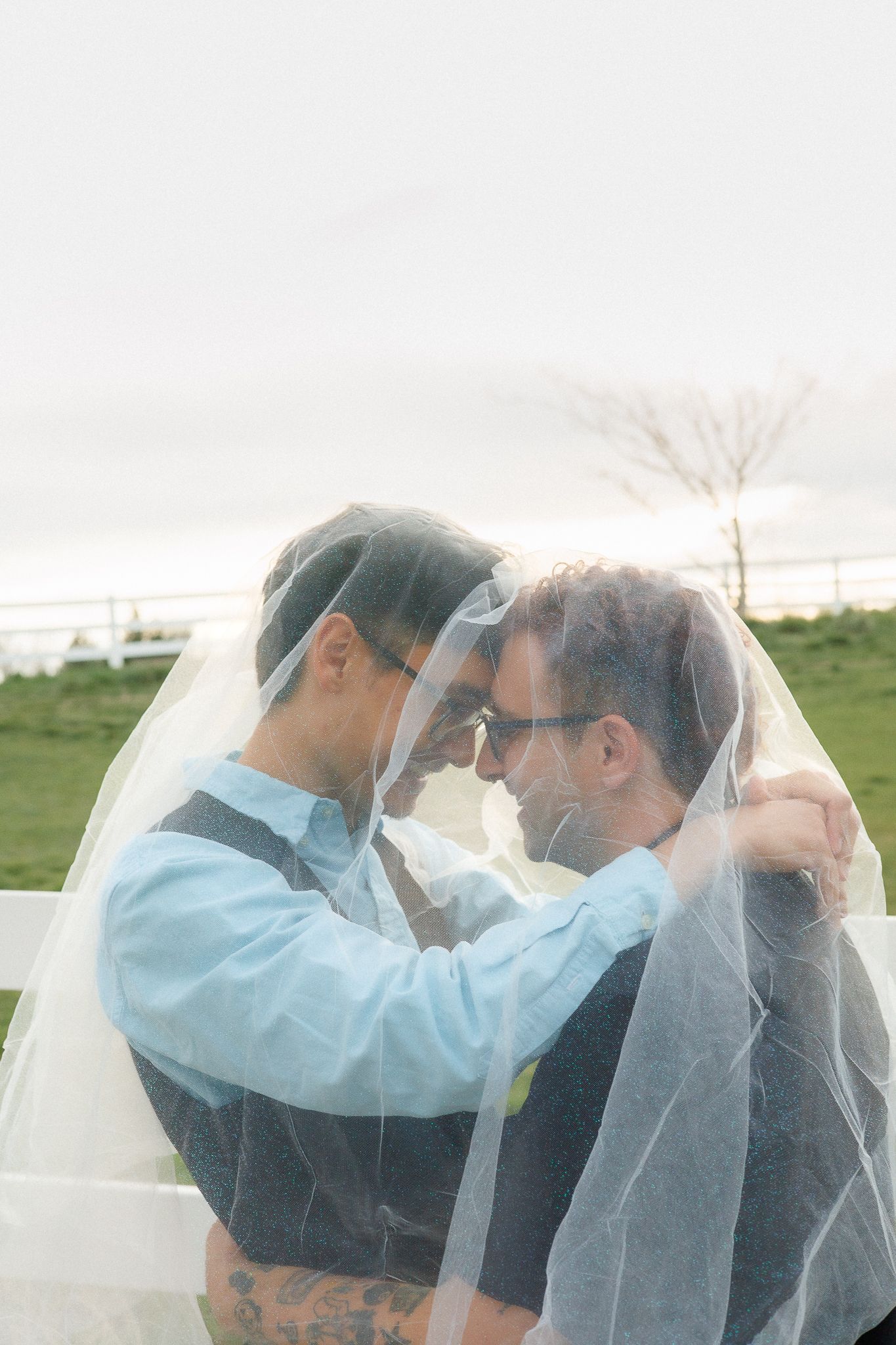 Two grooms share a romantic moment under a flowing white wedding veil at an outdoor ceremony.