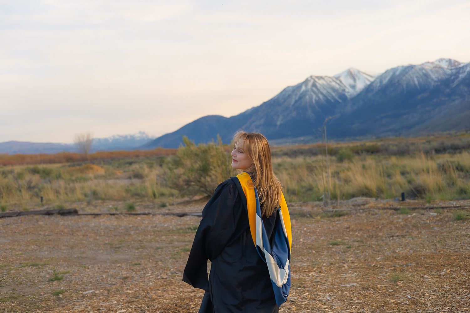 Young woman in graduation cap and gown stands in an open field with snow-capped mountains at sunset.