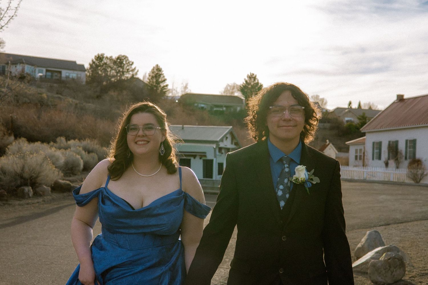 Young couple in prom attire holding hands outdoors, woman in blue off-shoulder gown and man in black suit with boutonniere.