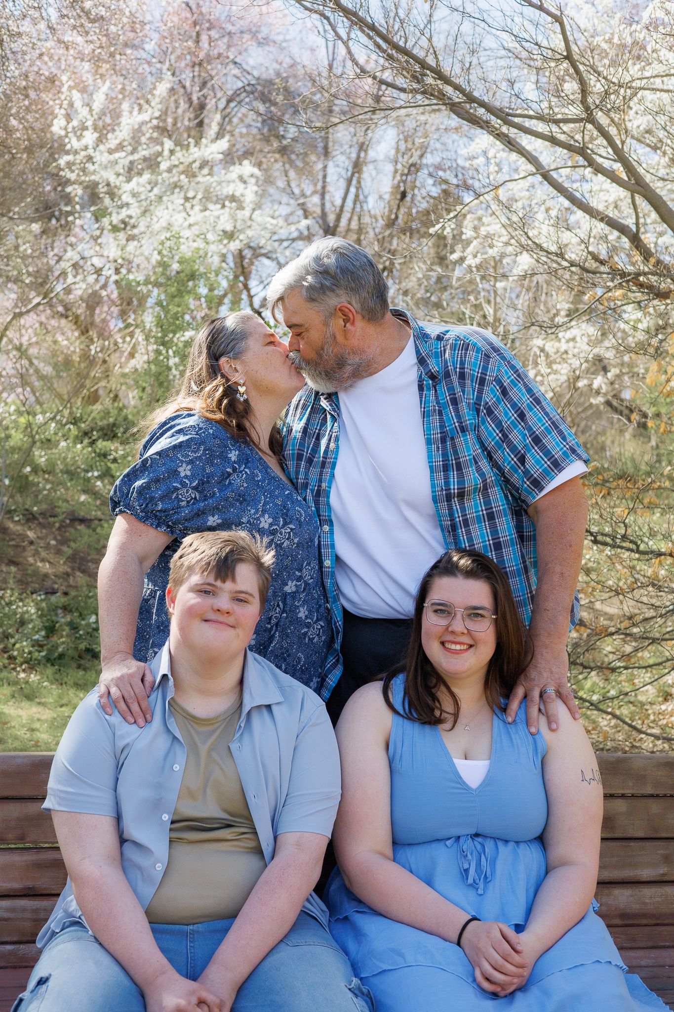 Parents kissing behind their two adult children seated on a bench surrounded by blooming spring trees.