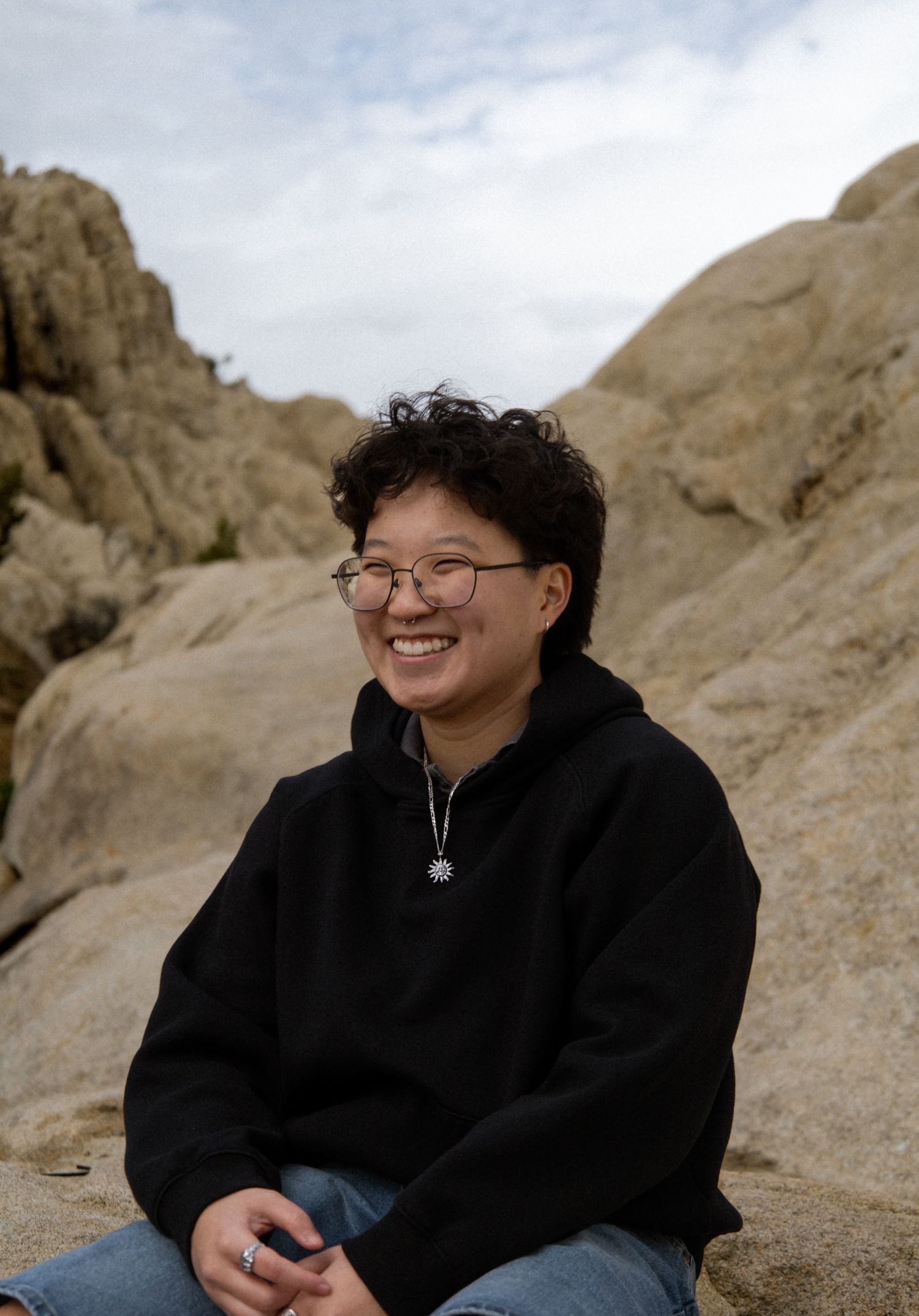 Smiling young person with curly hair in black hoodie sitting among boulders under cloudy sky.