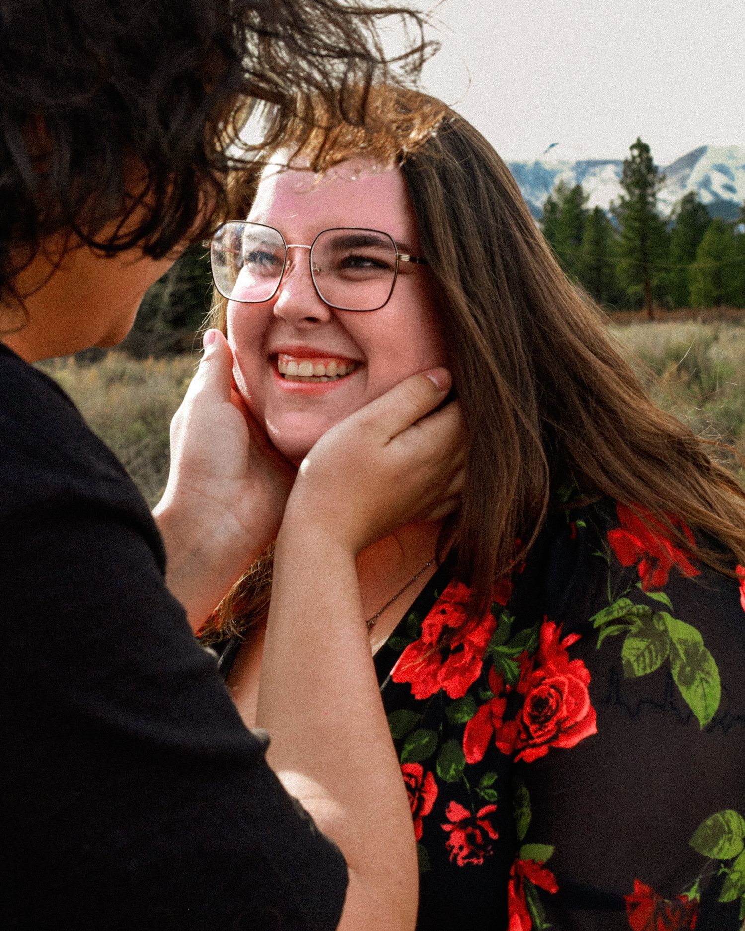 Young woman smiling warmly as her partner holds her face in a scenic mountain meadow setting.