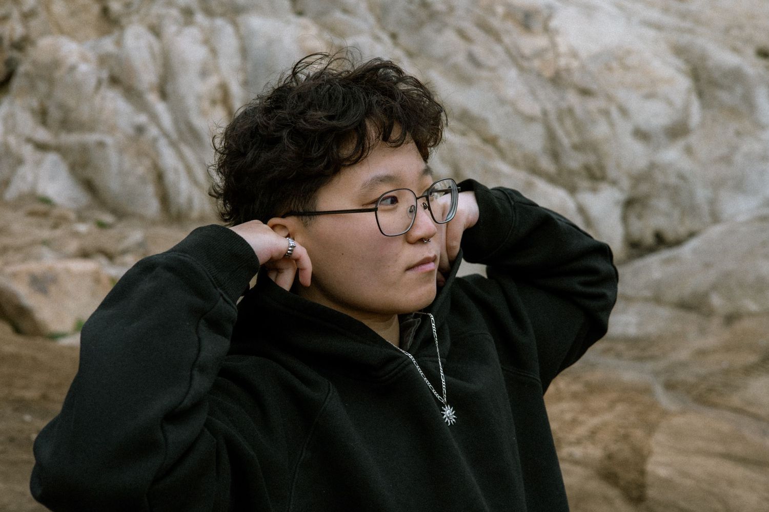 Young person in black hoodie adjusting glasses while sitting among large boulders outdoors.