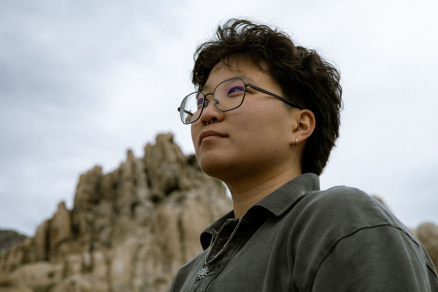 Young person with curly hair and glasses looking up against rocky landscape and cloudy sky.
