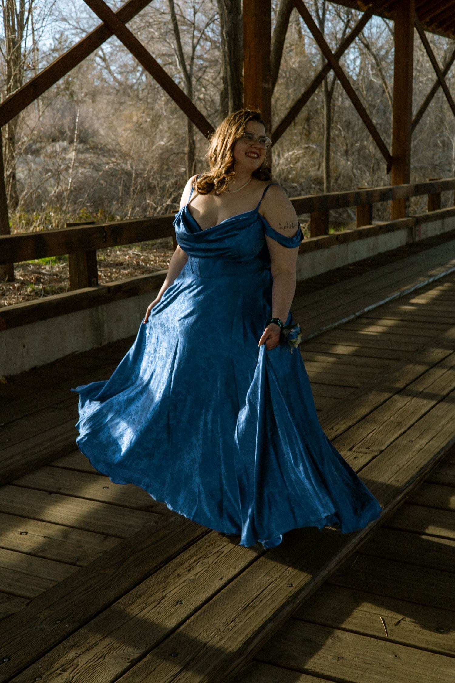 A woman in a flowing blue off-shoulder gown twirls on a wooden bridge in a forest.