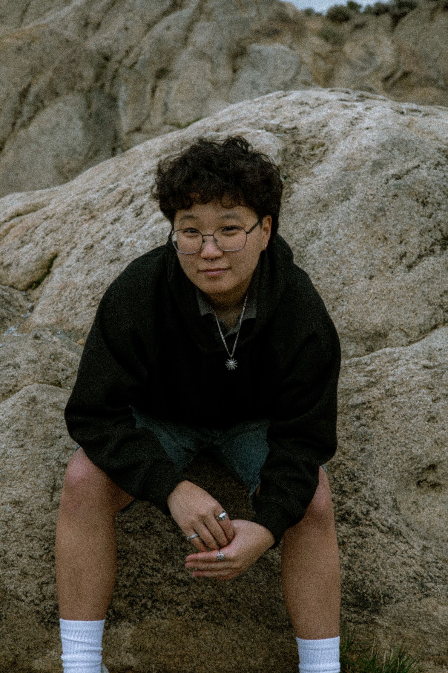 Young person with curly hair and glasses sitting on rocks outdoors in black hoodie and shorts.