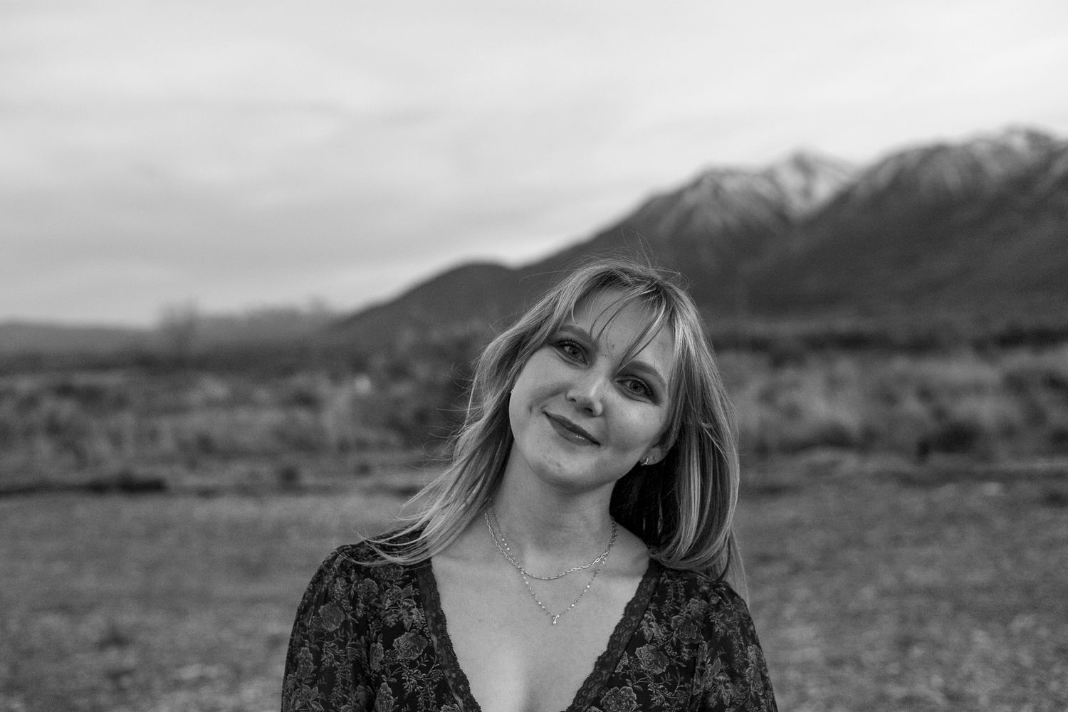 Black and white portrait of a smiling blonde woman outdoors with mountain landscape in background.