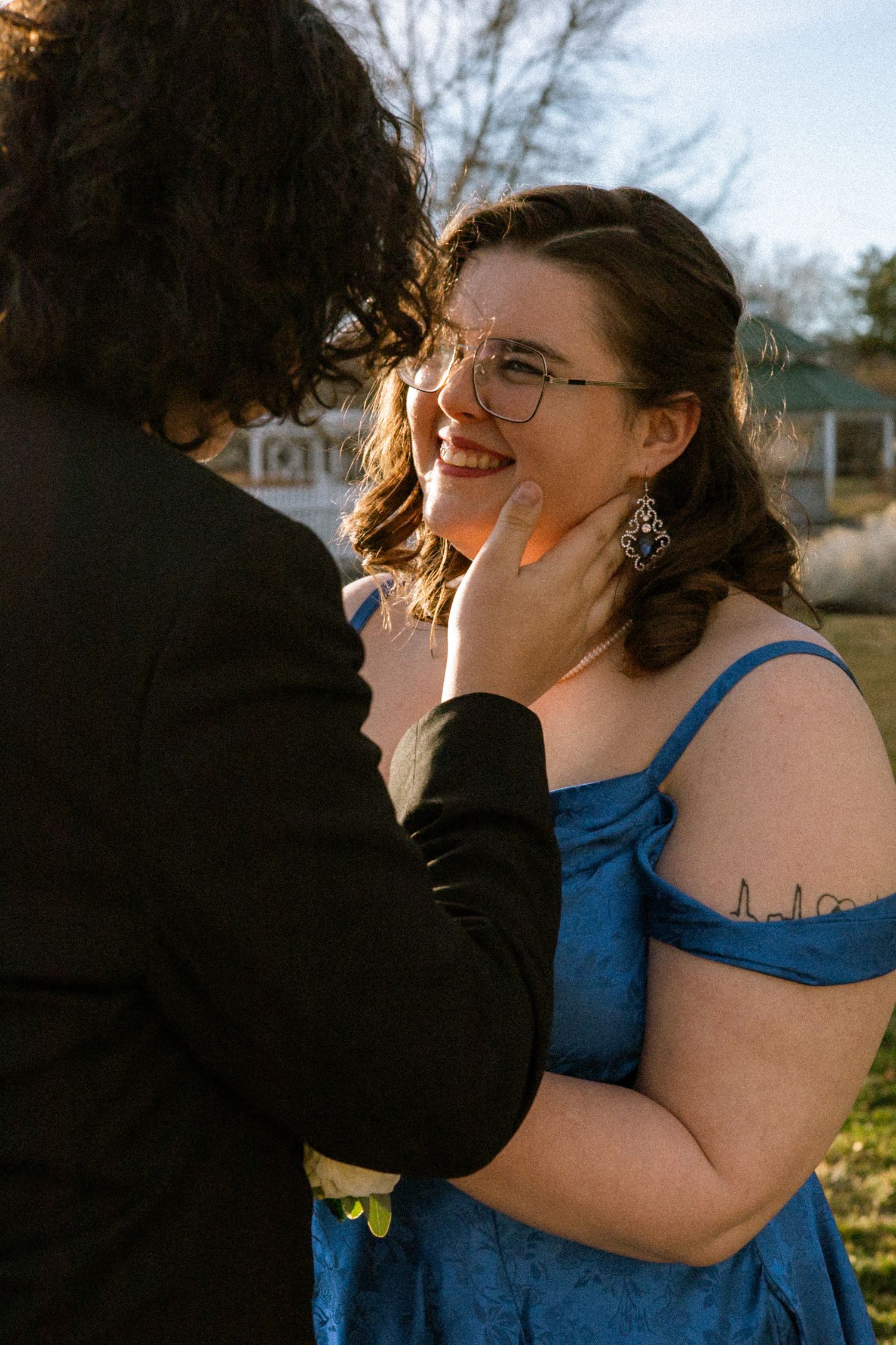 Young woman in blue prom dress smiling joyfully as her date gently touches her face outdoors.