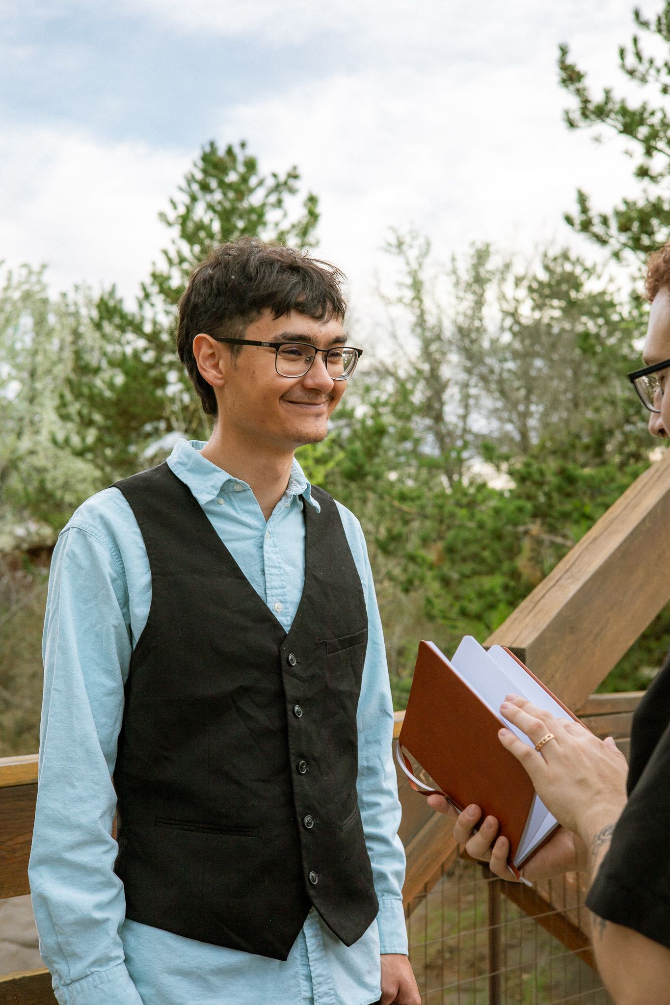 A smiling person in a light blue shirt and black vest stands outdoors during a wedding ceremony.