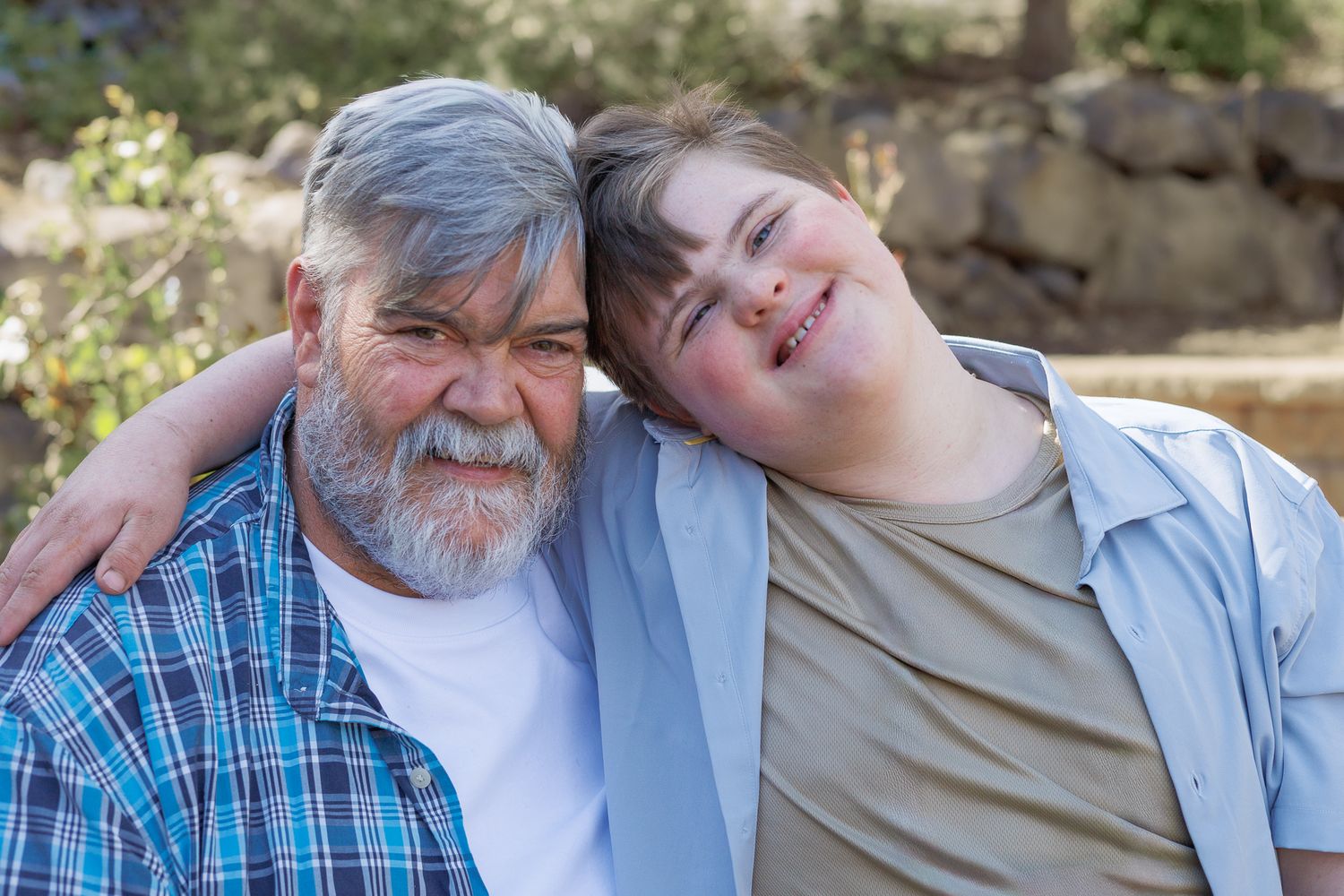 Older man with gray beard and teenage boy leaning on his shoulder smiling outdoors.
