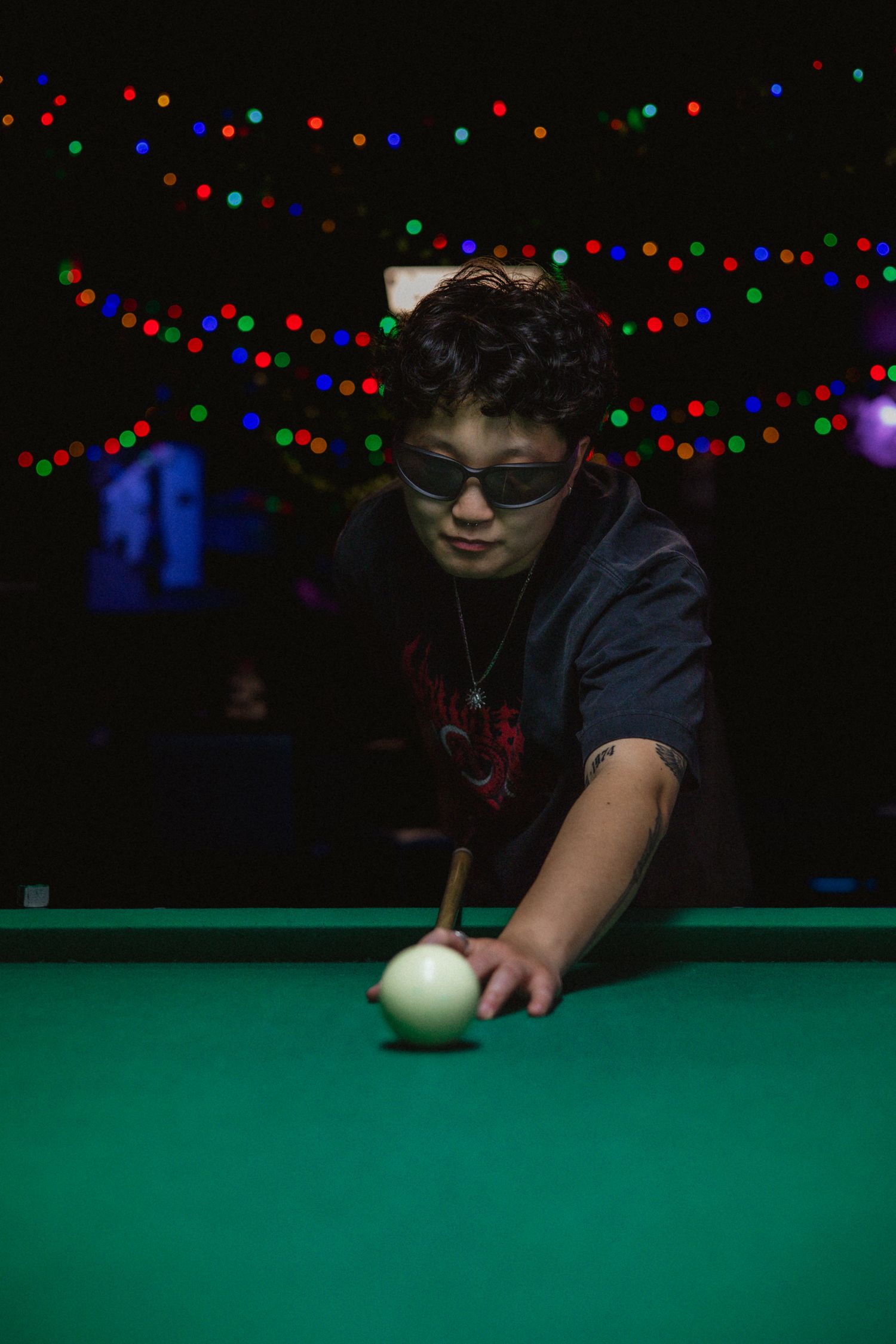 Young person in sunglasses leans over a green pool table to take a shot under colorful bokeh lights.
