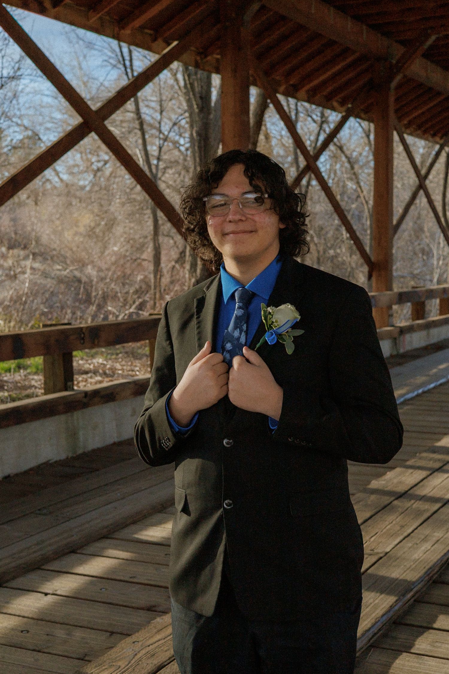 Smiling teenage boy in a black suit with blue shirt and boutonniere stands on a wooden bridge.