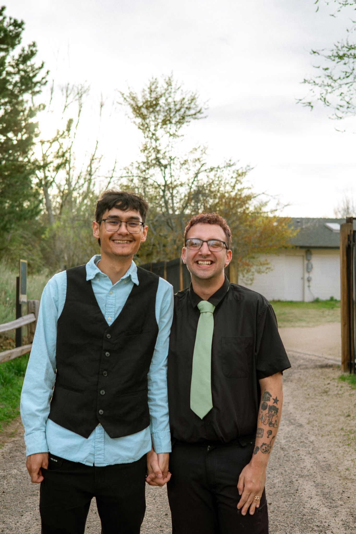 Two smiling men in formal attire holding hands outdoors, one wearing a black vest and the other a green tie.