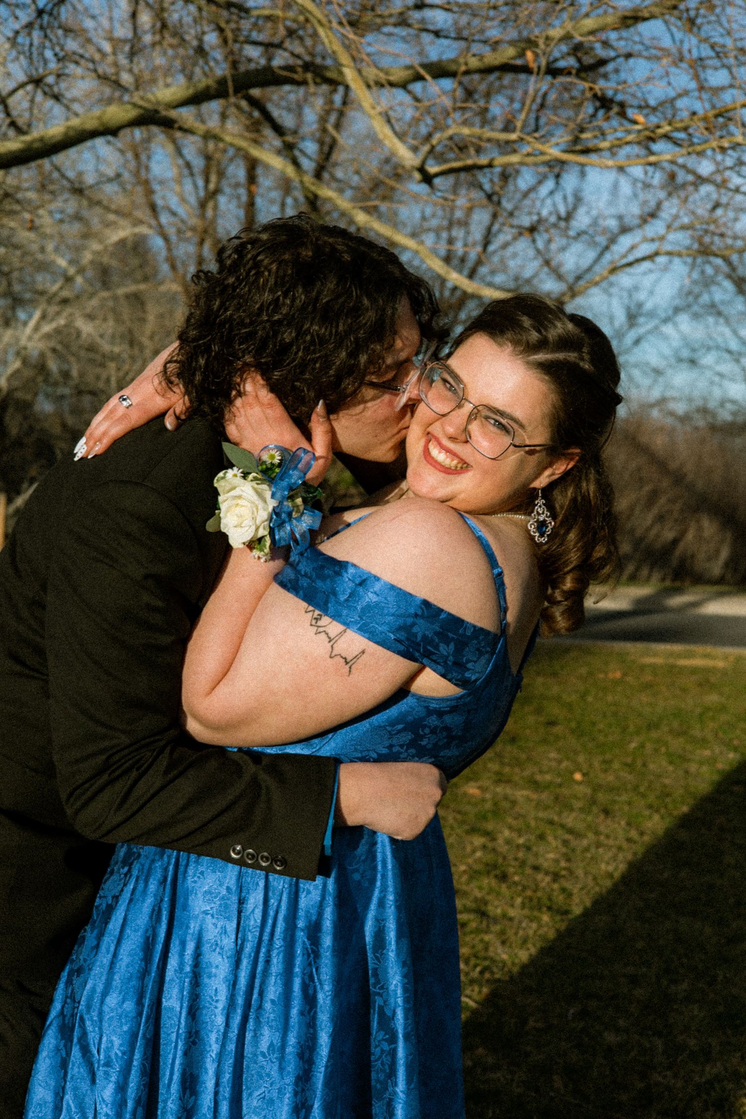 Prom couple embracing outdoors, young woman in blue gown smiling as her date kisses her cheek.