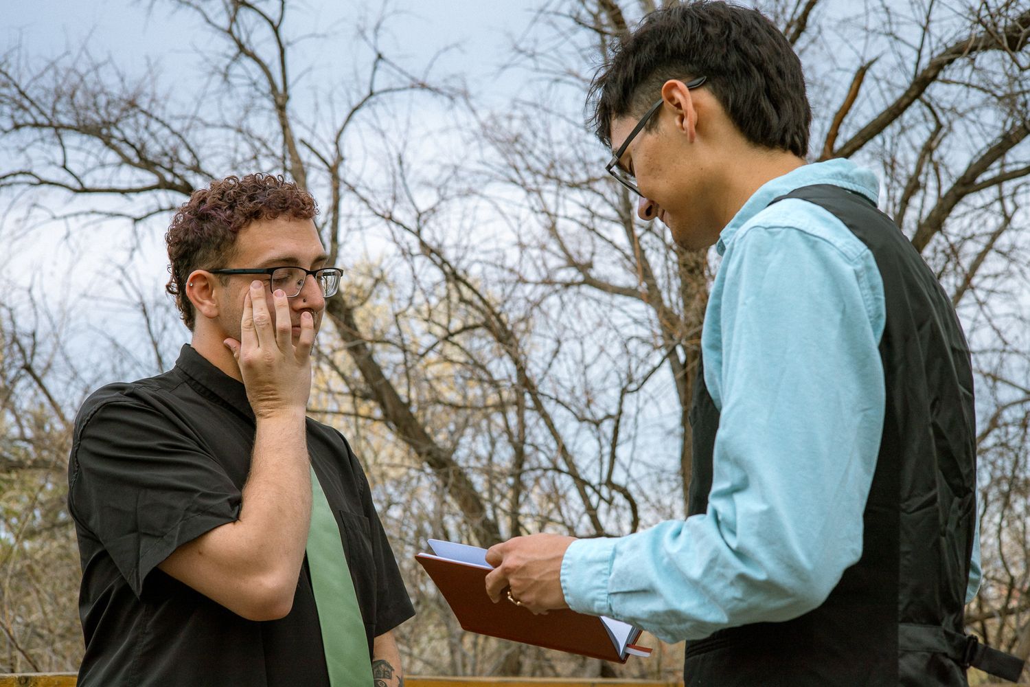 An emotional man wipes tears during an outdoor wedding ceremony while his partner reads vows.