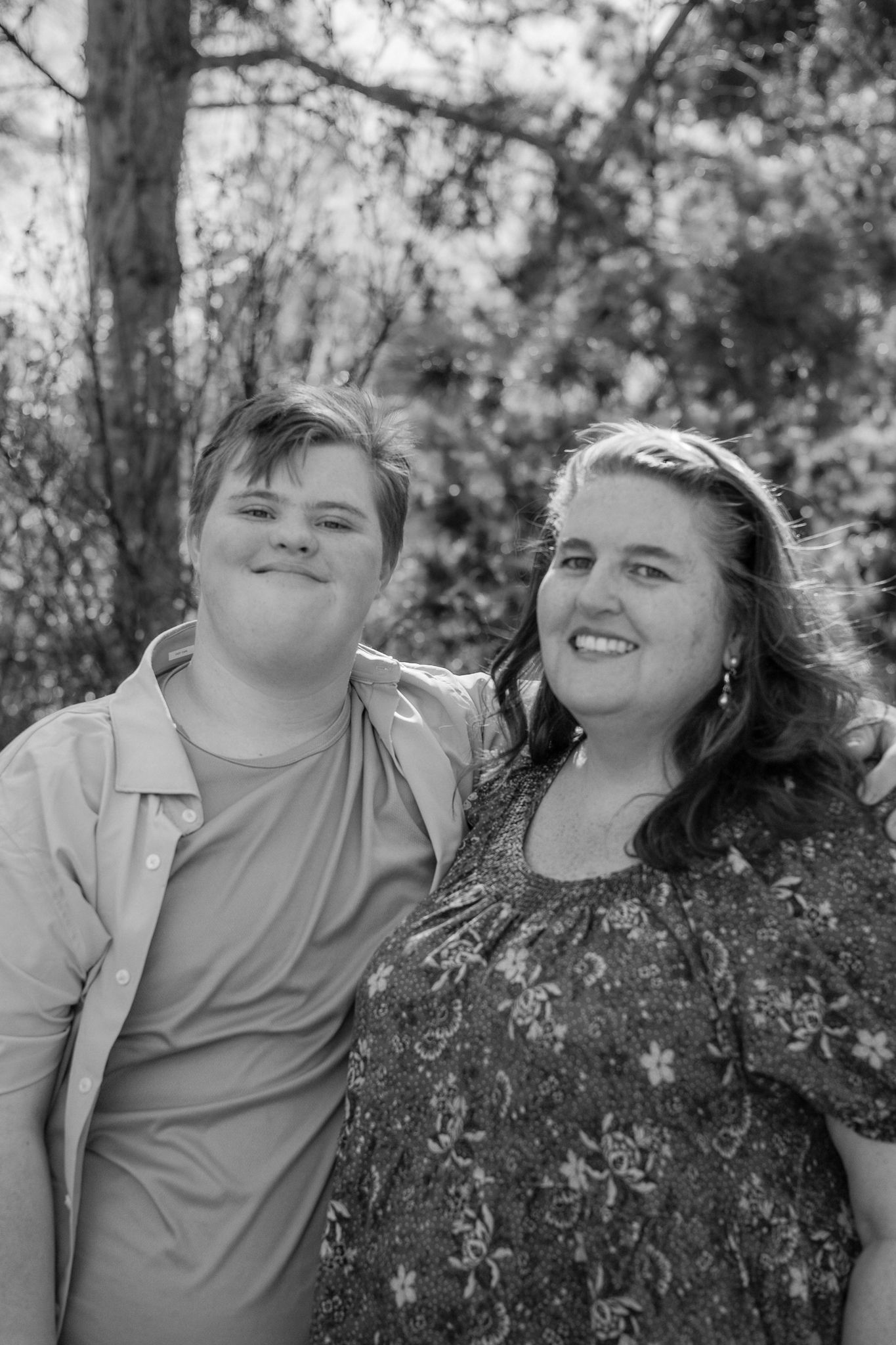 Black and white photo of a teenage boy and smiling woman posing together outdoors among trees.