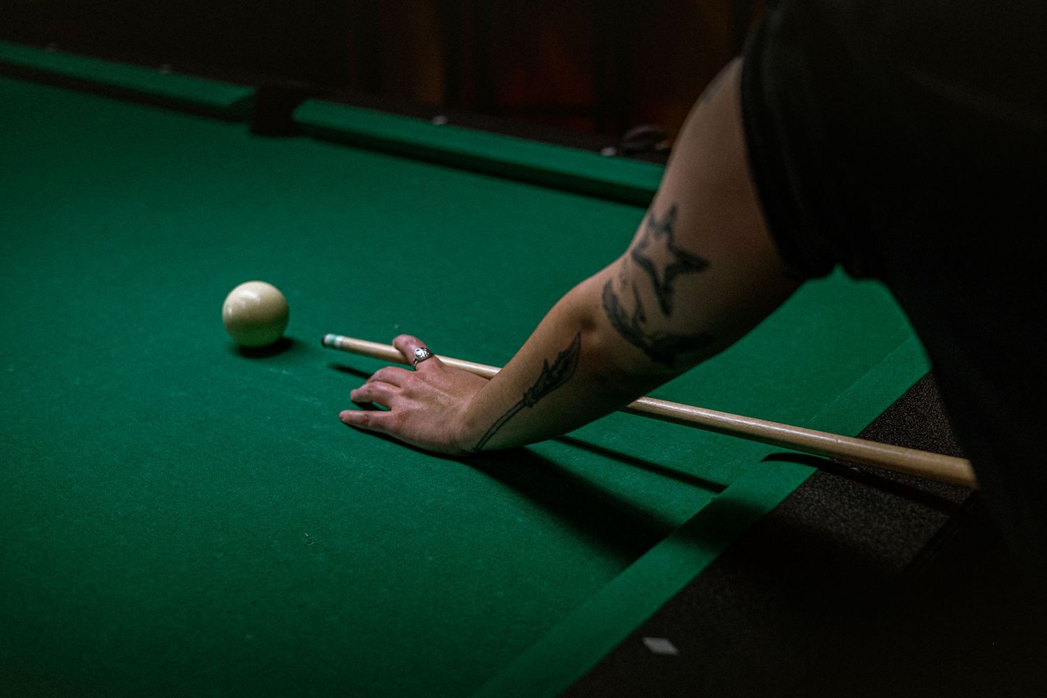 Close-up of tattooed hand positioned on green felt pool table holding a cue stick near the cue ball.