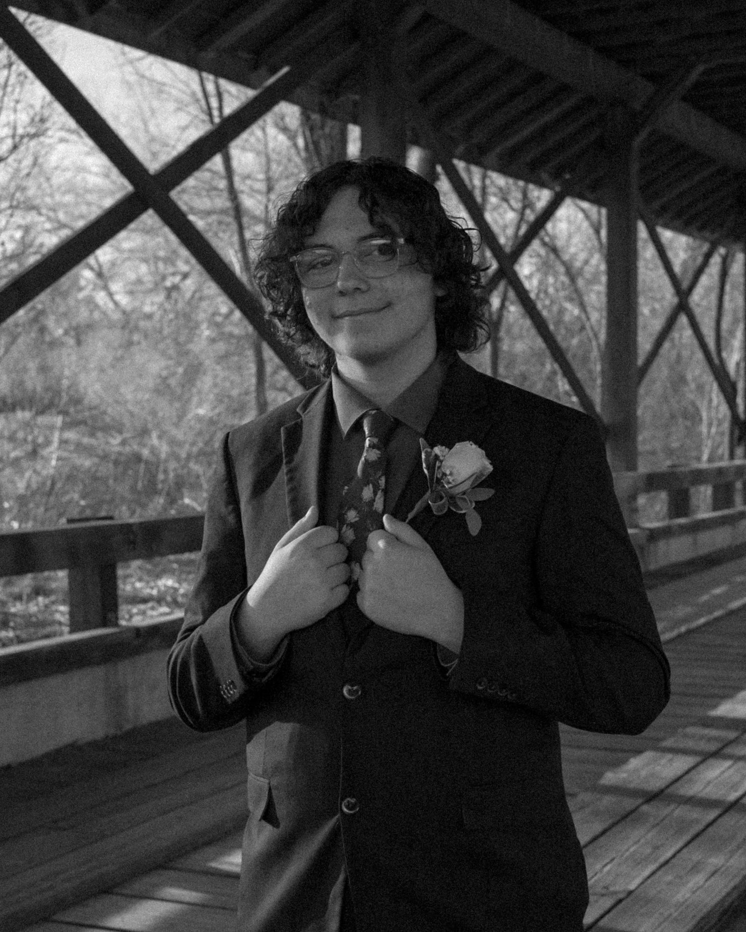 Black and white portrait of a teenage boy in a suit with boutonniere posing on a wooden bridge.
