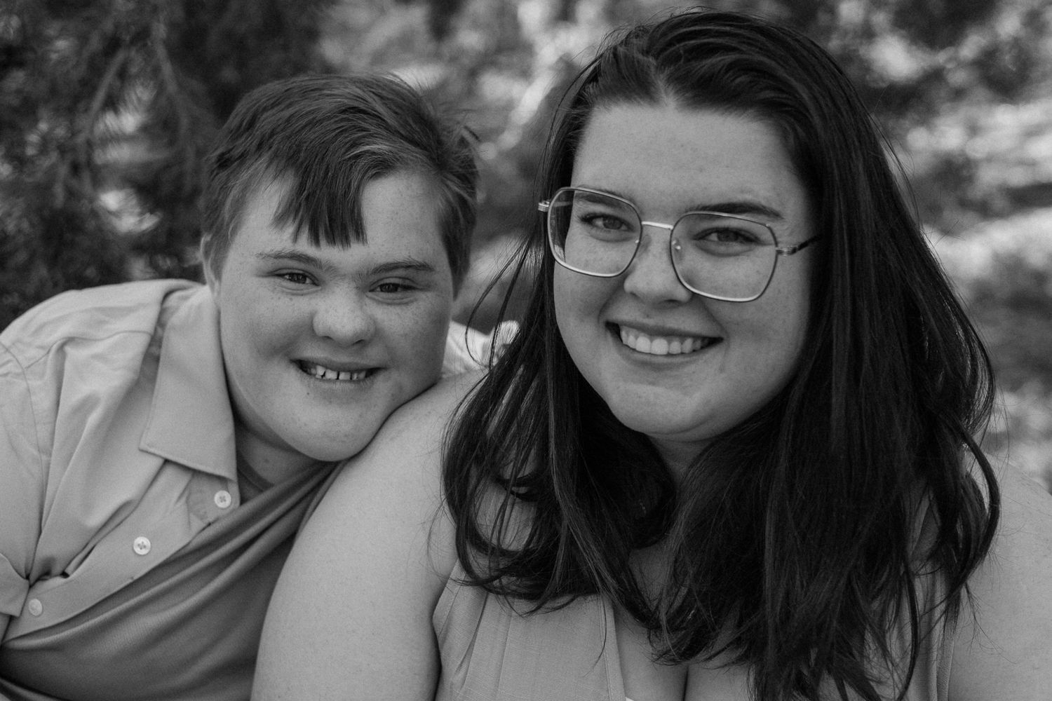 Black and white portrait of a young boy with Down syndrome and a teenage girl smiling together outdoors.