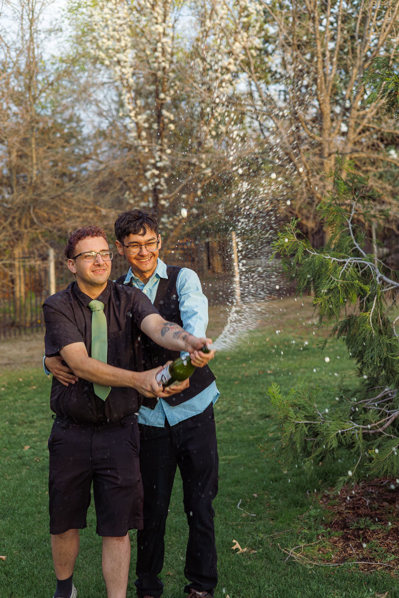 Two smiling men in formal wear pop champagne together in a blooming spring garden.