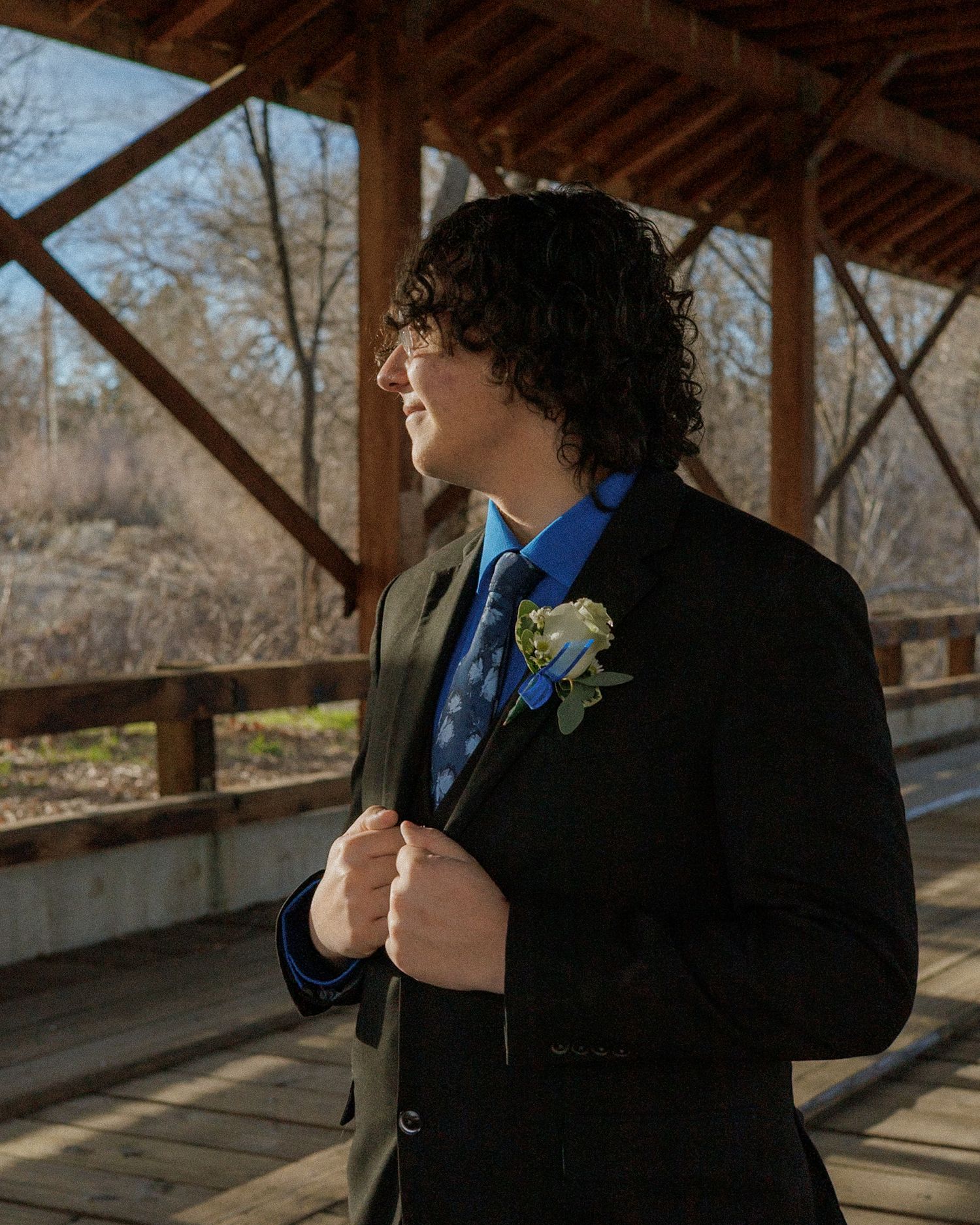 Teenage boy in a black suit with blue shirt and boutonniere poses on a wooden covered bridge.