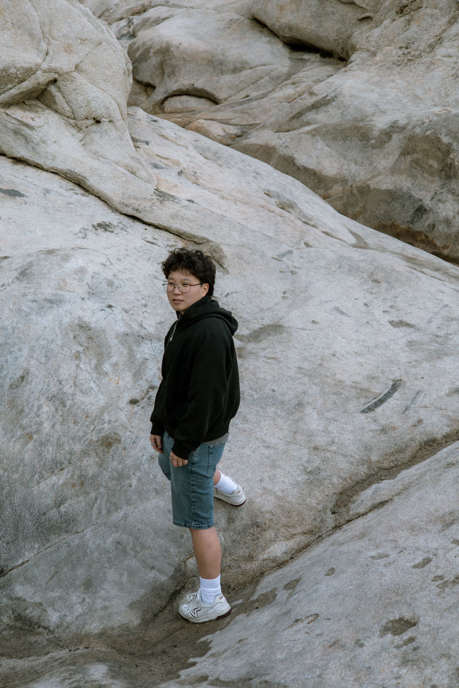 Young person in black hoodie and shorts standing on large rock formation, looking at camera.