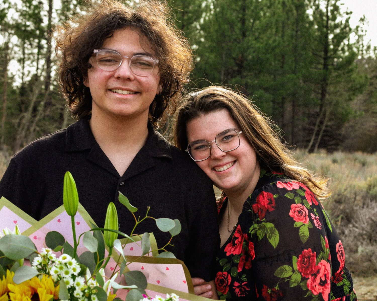 Smiling young couple holding a bouquet of flowers in a natural outdoor setting with pine trees.