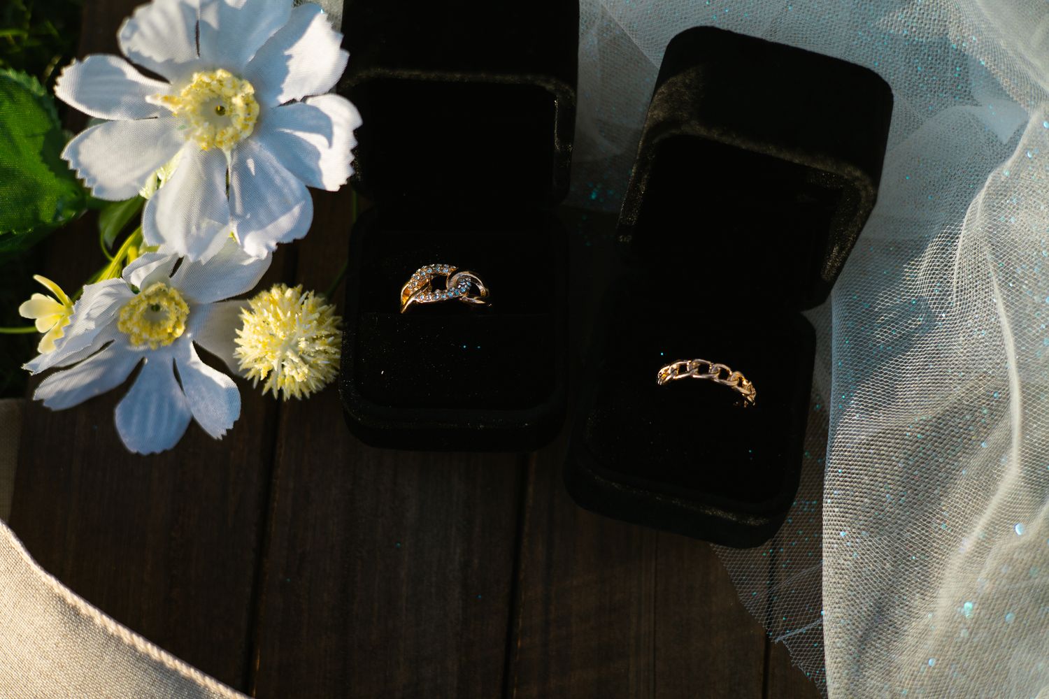 Two wedding rings in black velvet boxes displayed with white flowers and a sheer veil on wood.